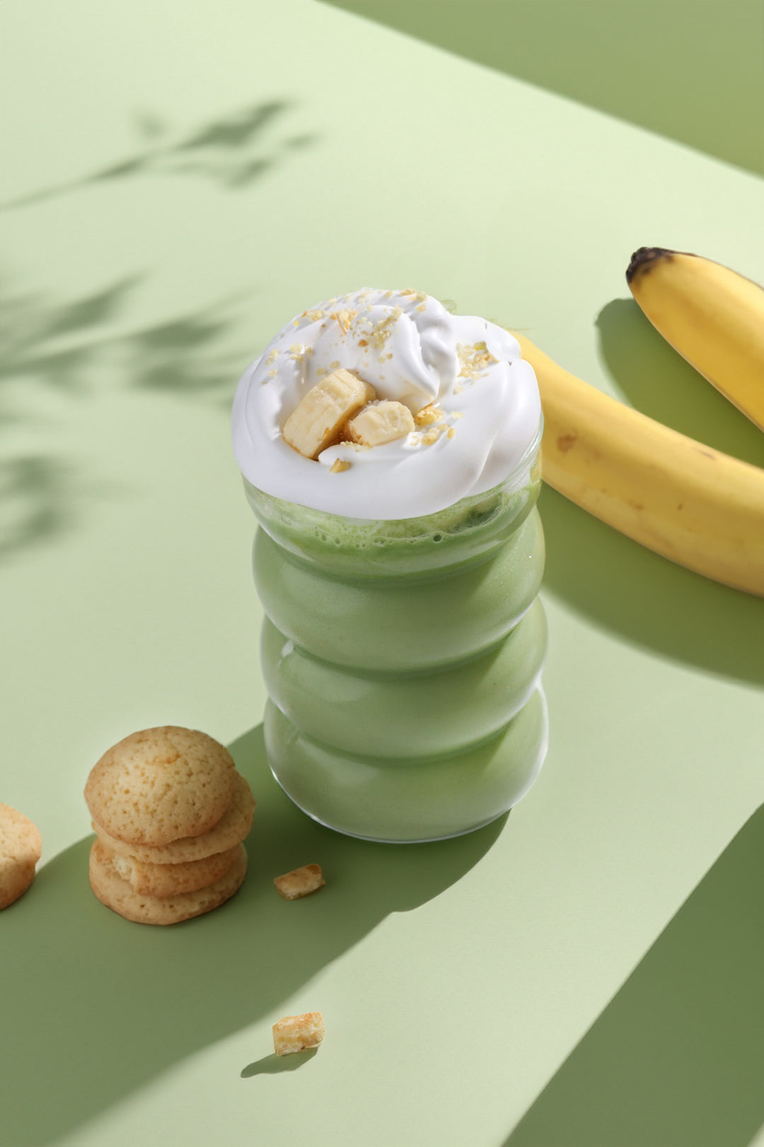A vibrant green banana pudding matcha latte topped with a generous swirl of whipped cream and banana slices, paired with a stack of golden Nilla wafer cookies and fresh bananas in the background.