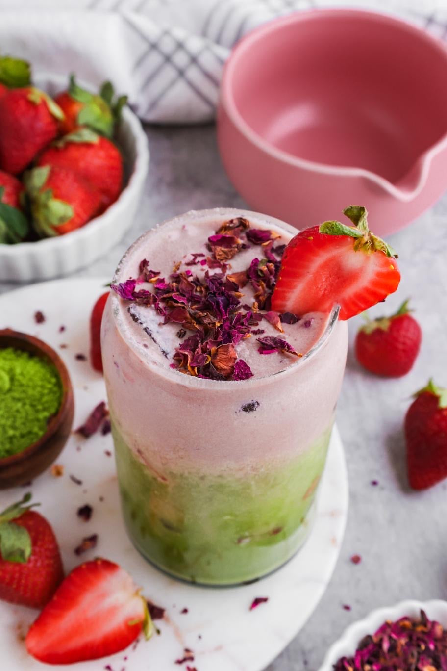 A refreshing strawberry rose iced matcha latte topped with vibrant rose petals and garnished with a fresh strawberry slice, surrounded by whole strawberries and a bowl of matcha powder, creating a visually appealing and delicious beverage scene.