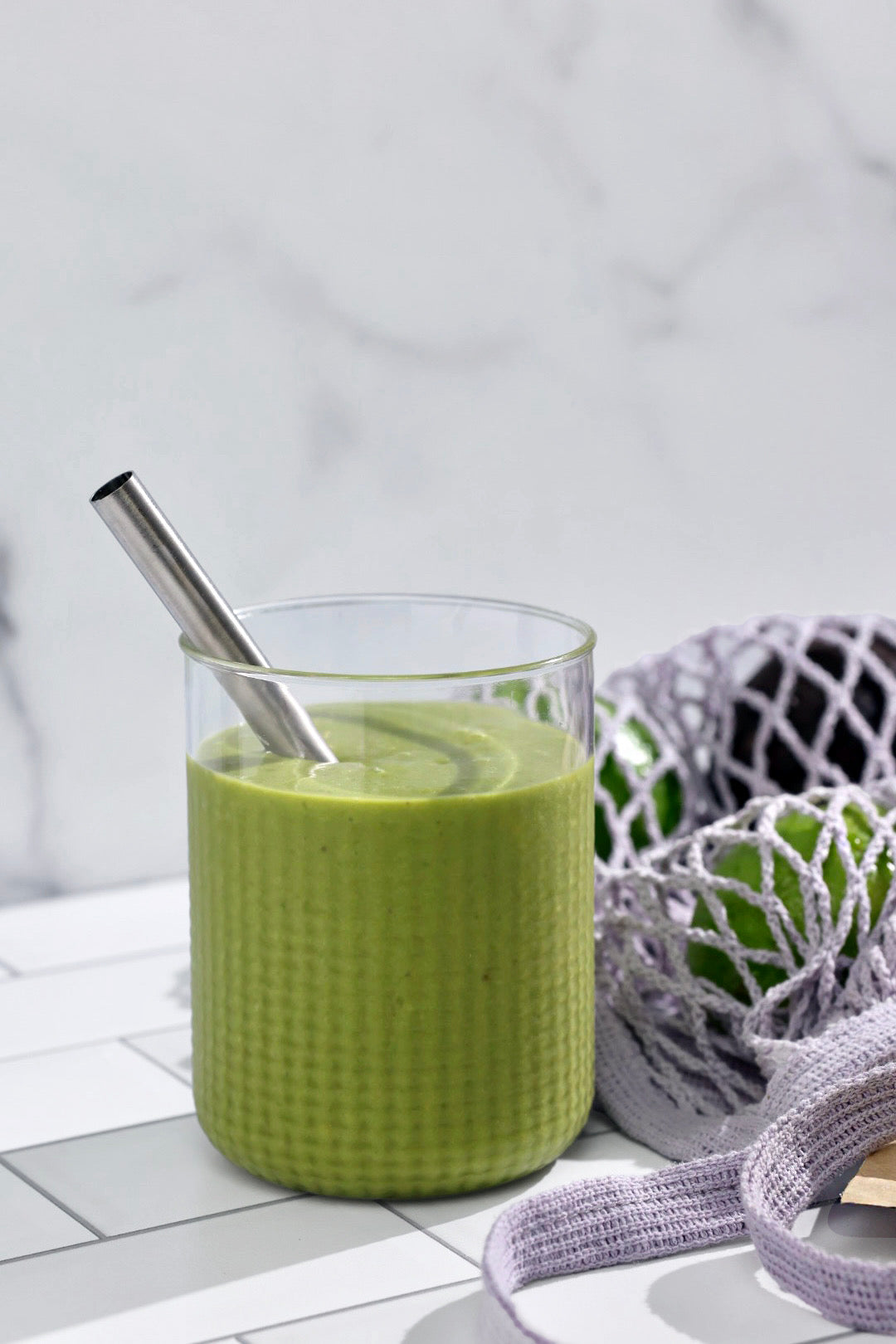 A creamy green matcha smoothie in a textured glass with a stainless steel straw, set on a tiled countertop beside a reusable mesh produce bag filled with green fruits.