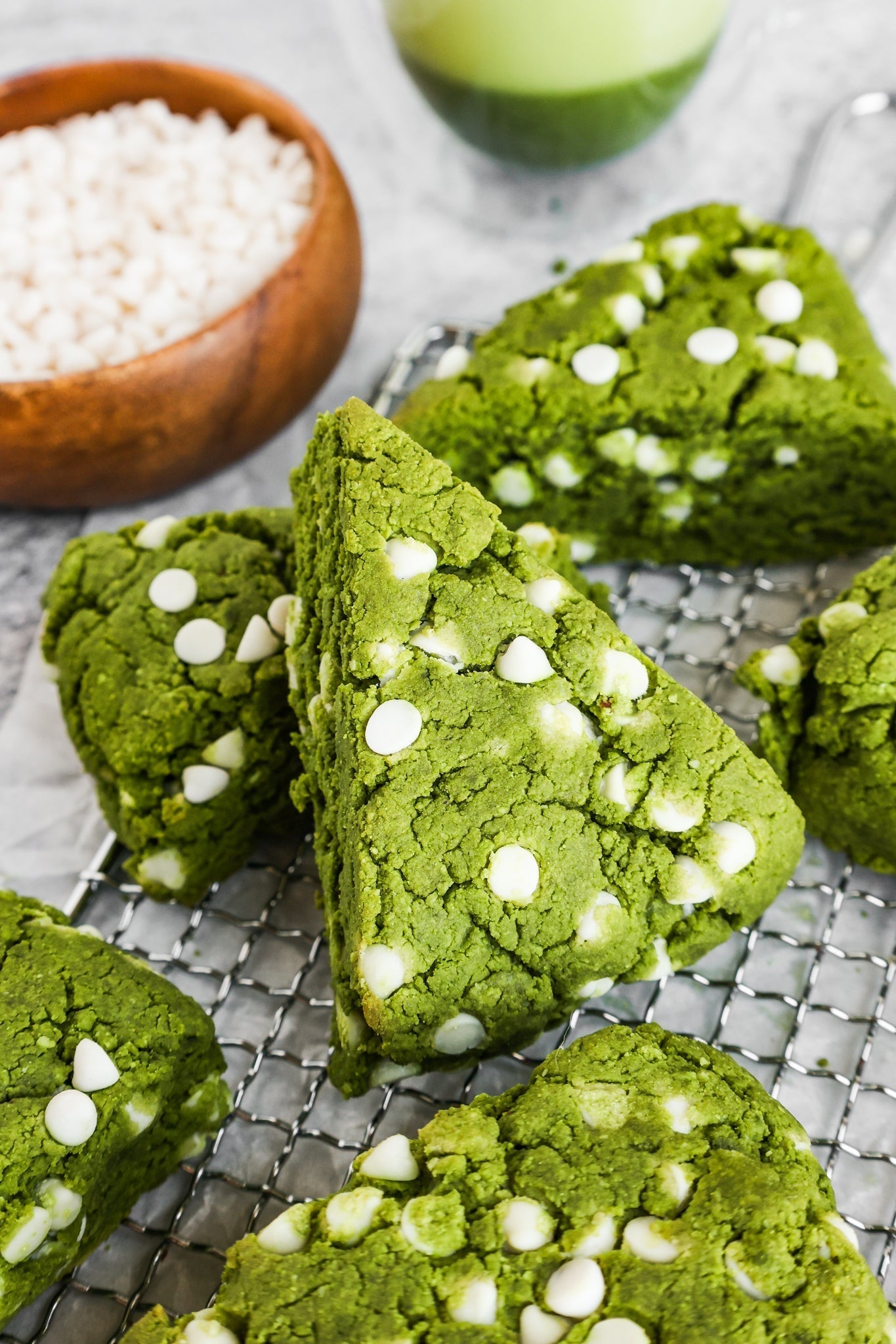 Freshly baked green matcha scones with white chocolate chips resting on a cooling rack, surrounded by a wooden bowl of white chocolate chips and a vibrant green matcha latte in the background.