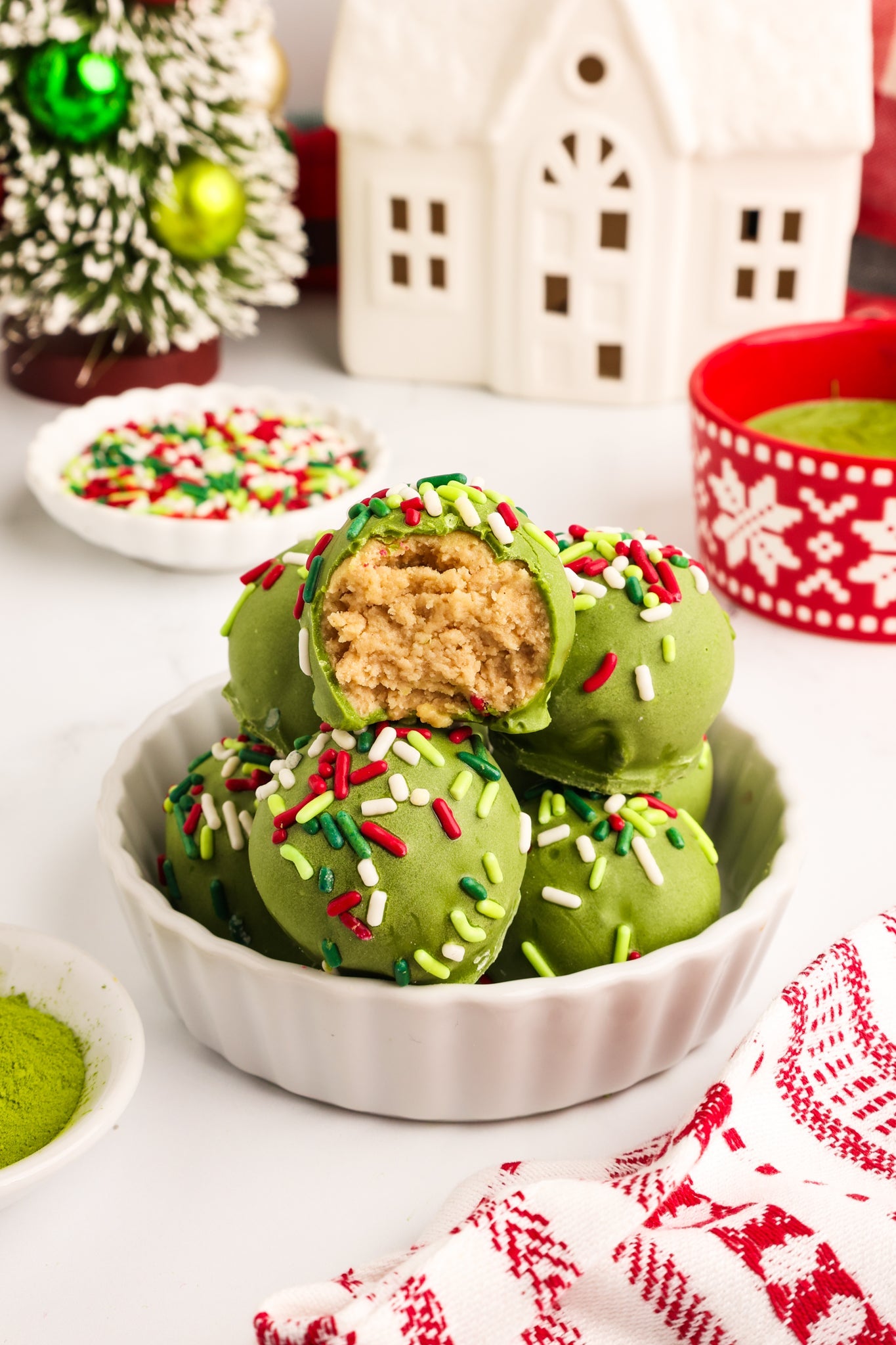 A festive plate of Matcha Sugar Cookie Truffles, coated in vibrant green matcha chocolate and topped with colorful Christmas sprinkles. One truffle is cut open to reveal a soft, cookie-like interior, surrounded by cozy holiday decor and twinkling lights.