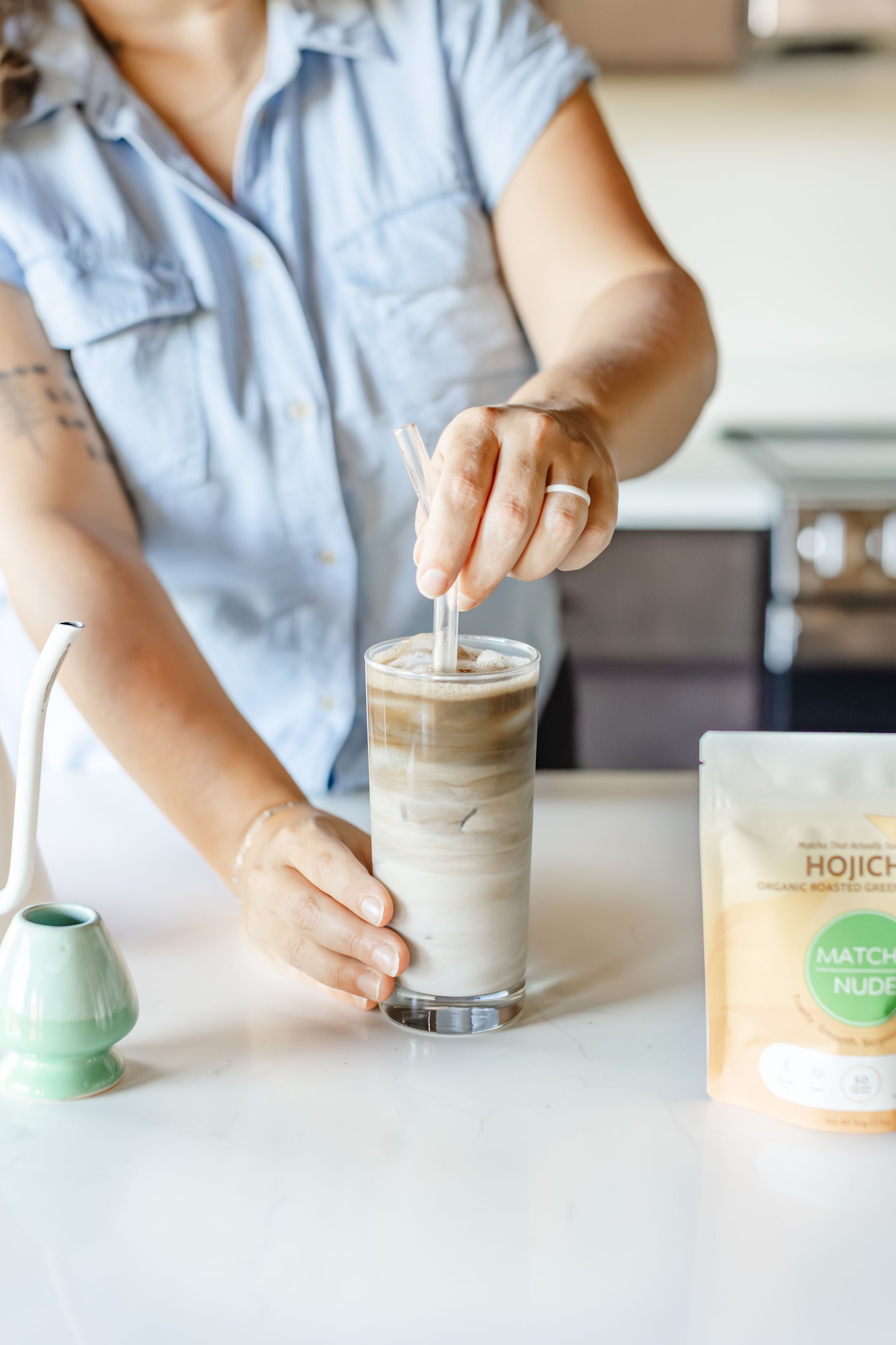Iced hojicha latte being stirred in a tall glass beside a bag of Matcha Nude organic hojicha and matcha-making tools.
