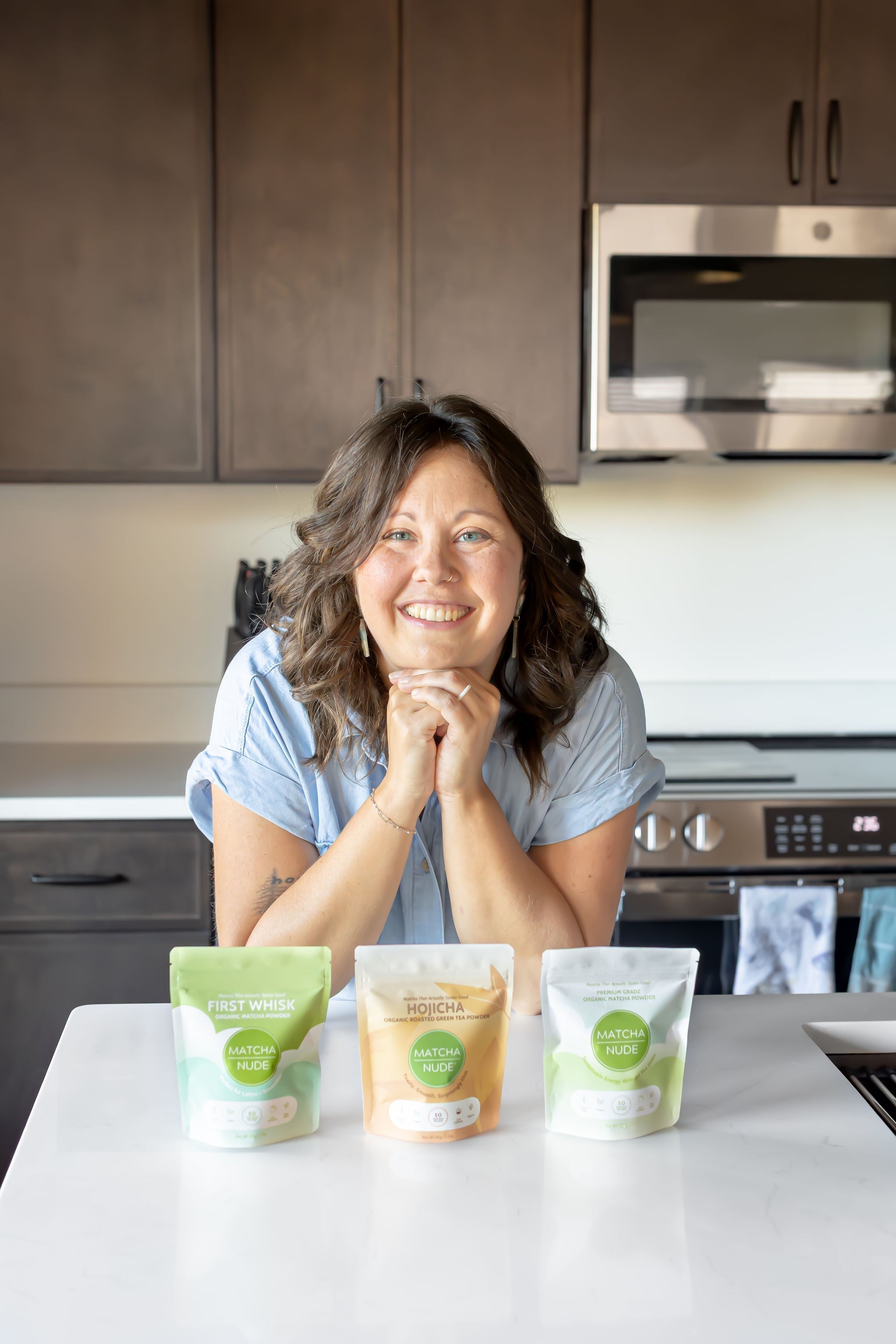 A smiling woman leans on a kitchen counter behind three Matcha Nude products: First Whisk matcha, Hojicha powder, and Premium matcha.