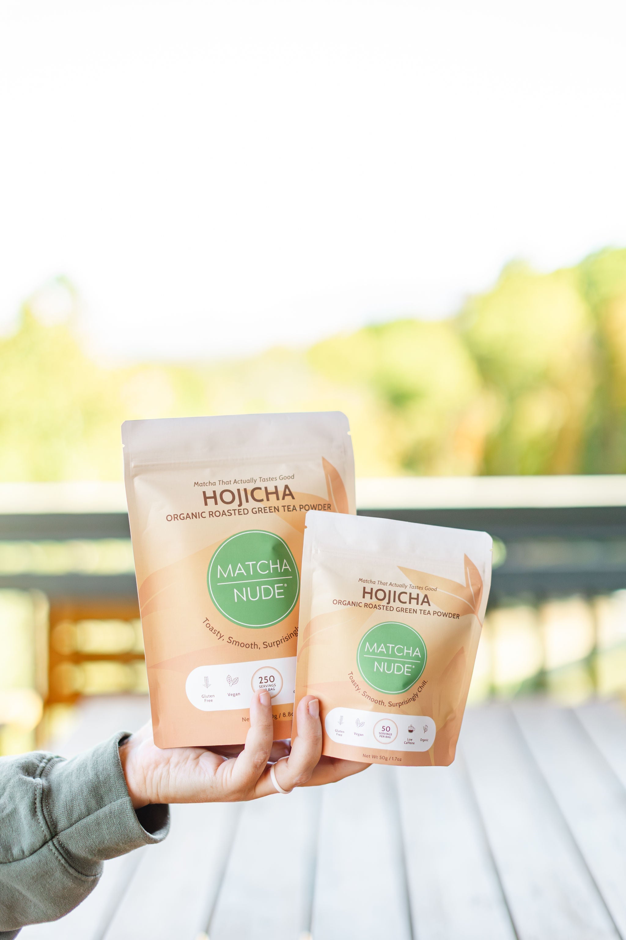 A woman in a green sweatshirt holding a 50g bag and a 250g bag of Matcha Nude hojicha