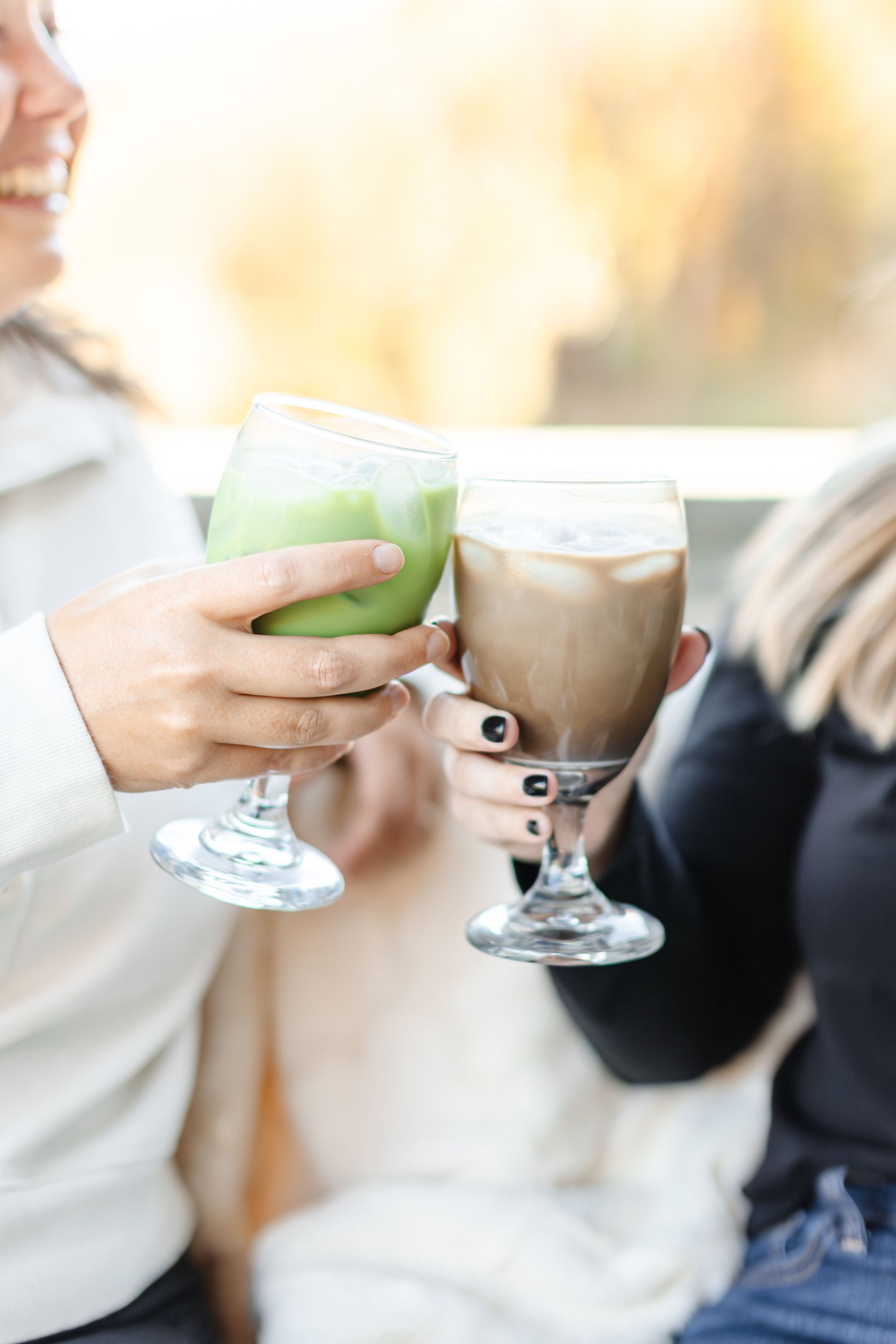 A smiling woman holding a matcha latte and another woman holding a hojicha latte made by Matcha Nude