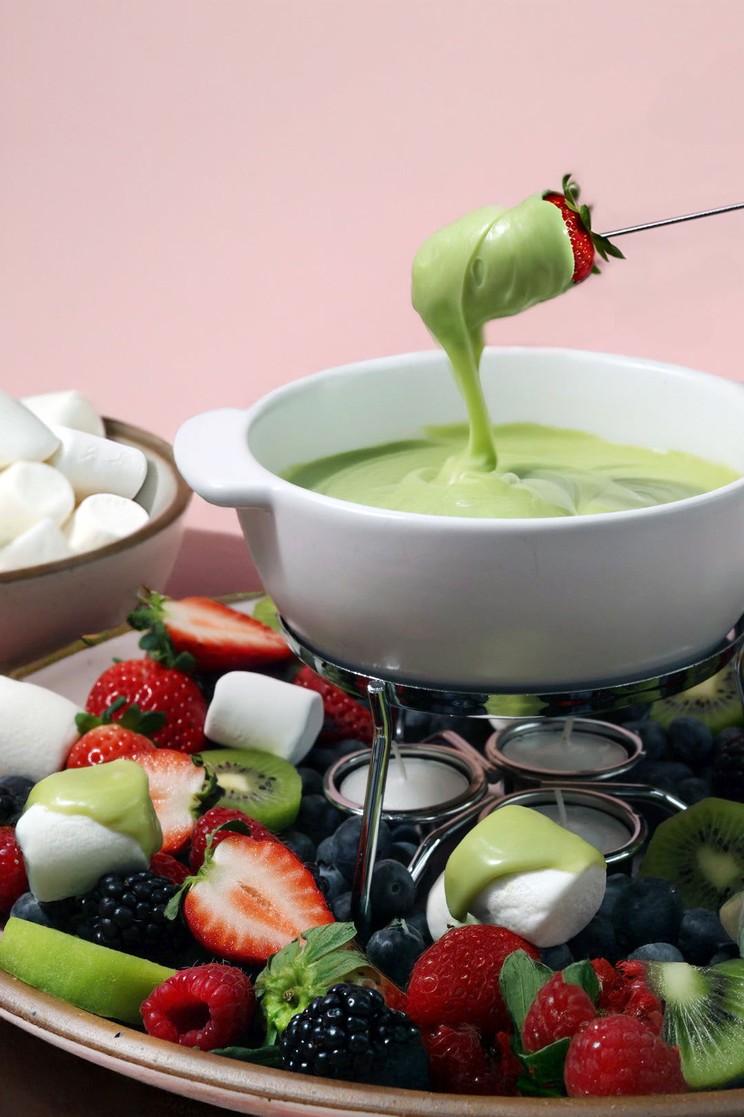 A delicious fondue setup featuring a white ceramic pot filled with creamy green matcha-flavored chocolate. A fresh strawberry is being dipped into the smooth, melted mixture.