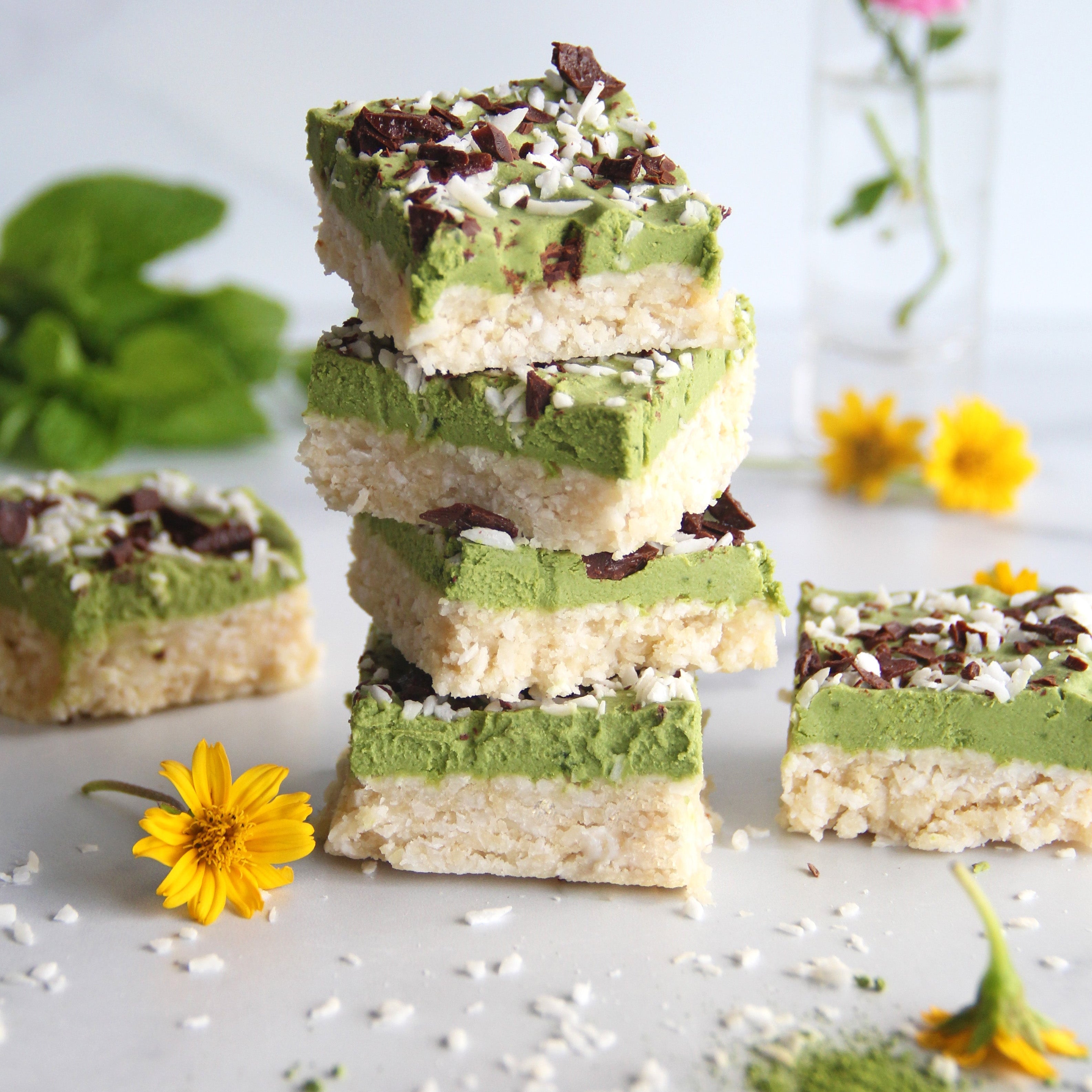 Stack of matcha mint coconut bars with a vibrant green layer, topped with shredded coconut and chocolate, surrounded by flowers and matcha powder.