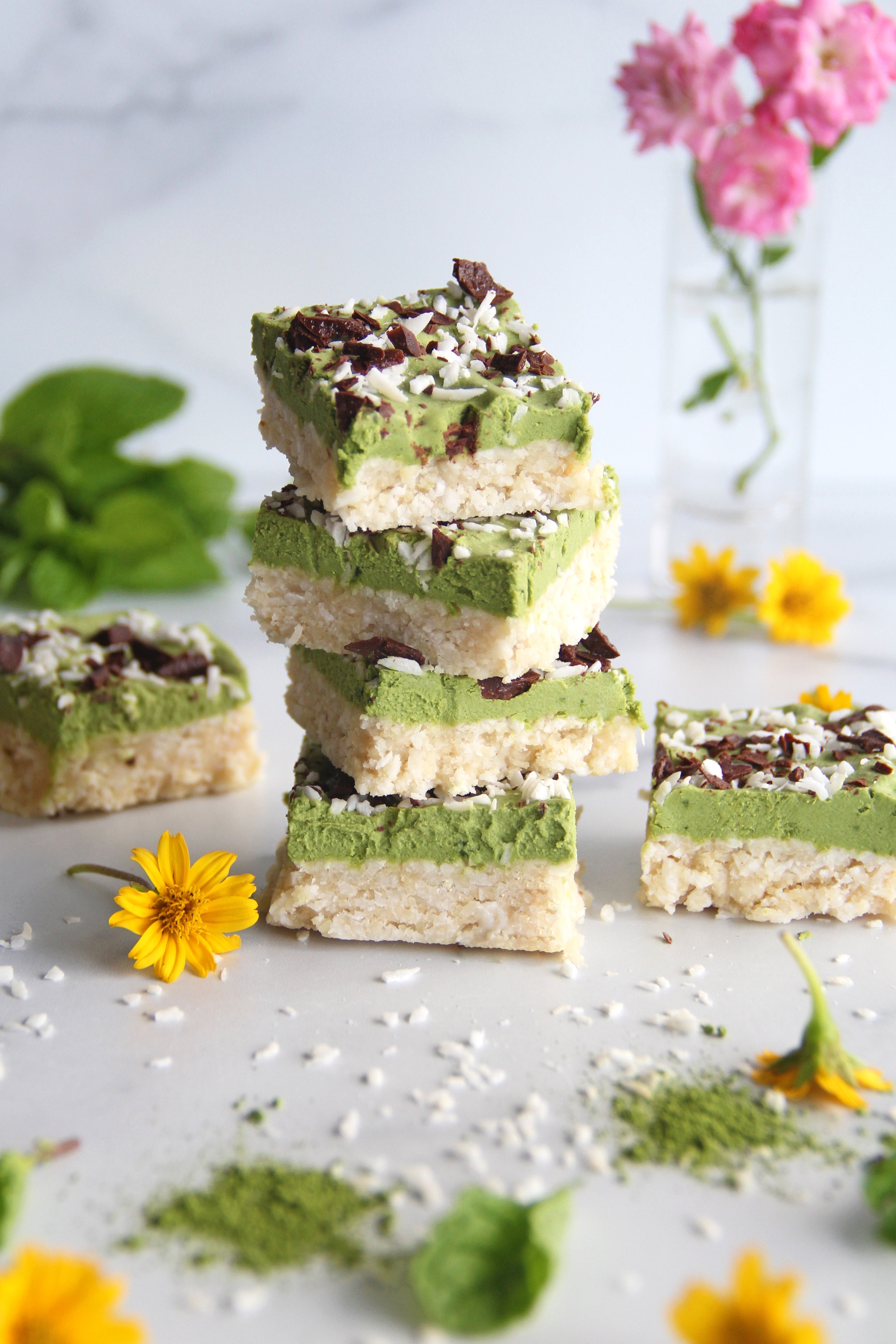 Stack of matcha mint coconut bars with a vibrant green layer, topped with shredded coconut and chocolate, surrounded by flowers and matcha powder.