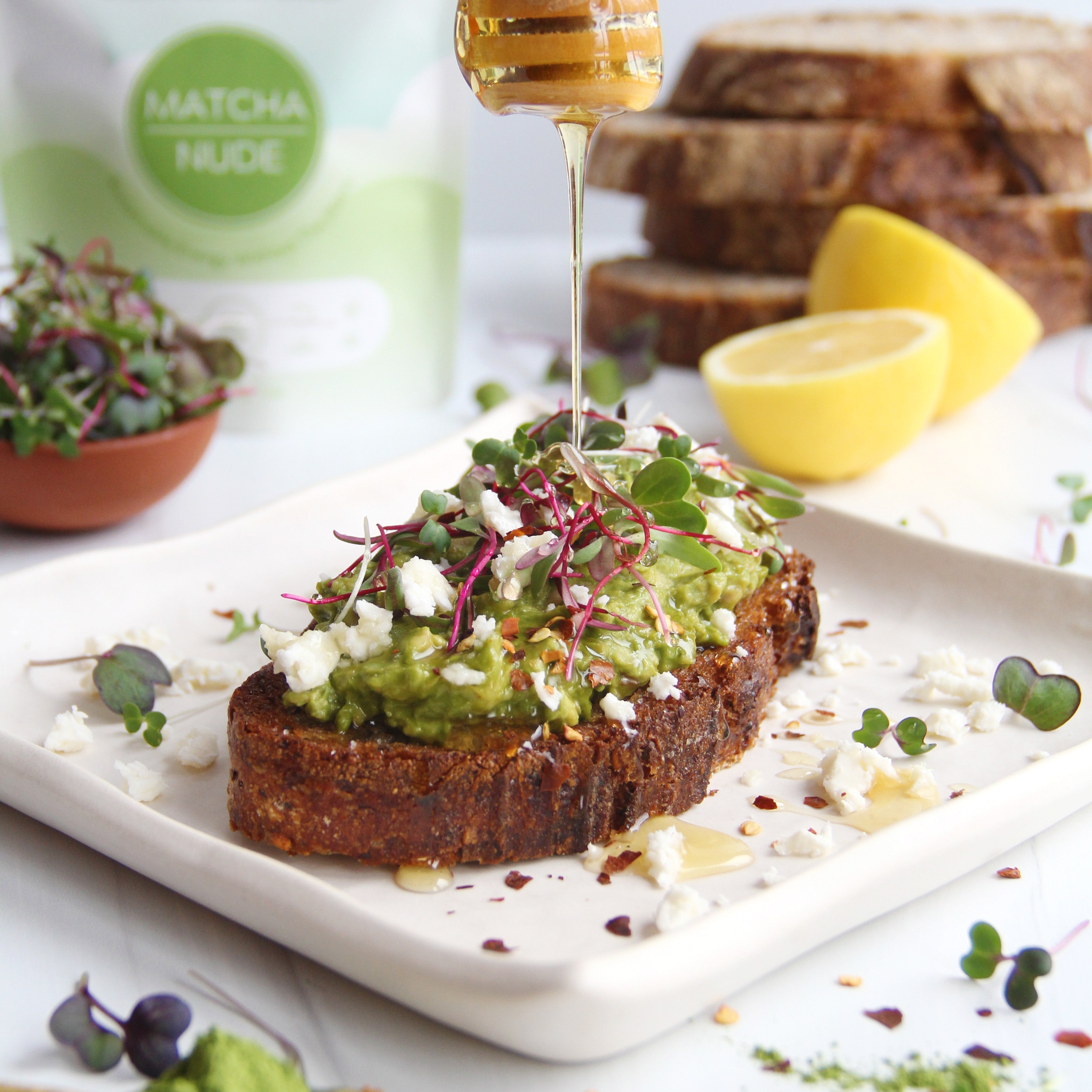 Matcha avocado toast topped with crumbled feta, microgreens, chili flakes, and a drizzle of honey on rustic bread, with Matcha Nude powder and lemon in the background.