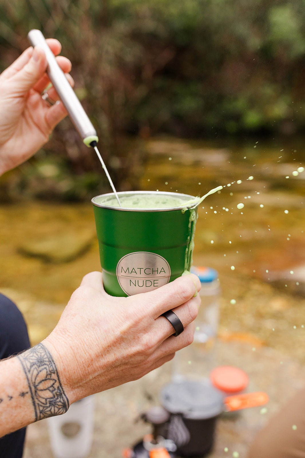 A close-up of a person frothing a vibrant green matcha latte in a green insulated Matcha Nude cup. The latte is frothy and overflowing slightly as it's being whisked, with drops of matcha splashing out.