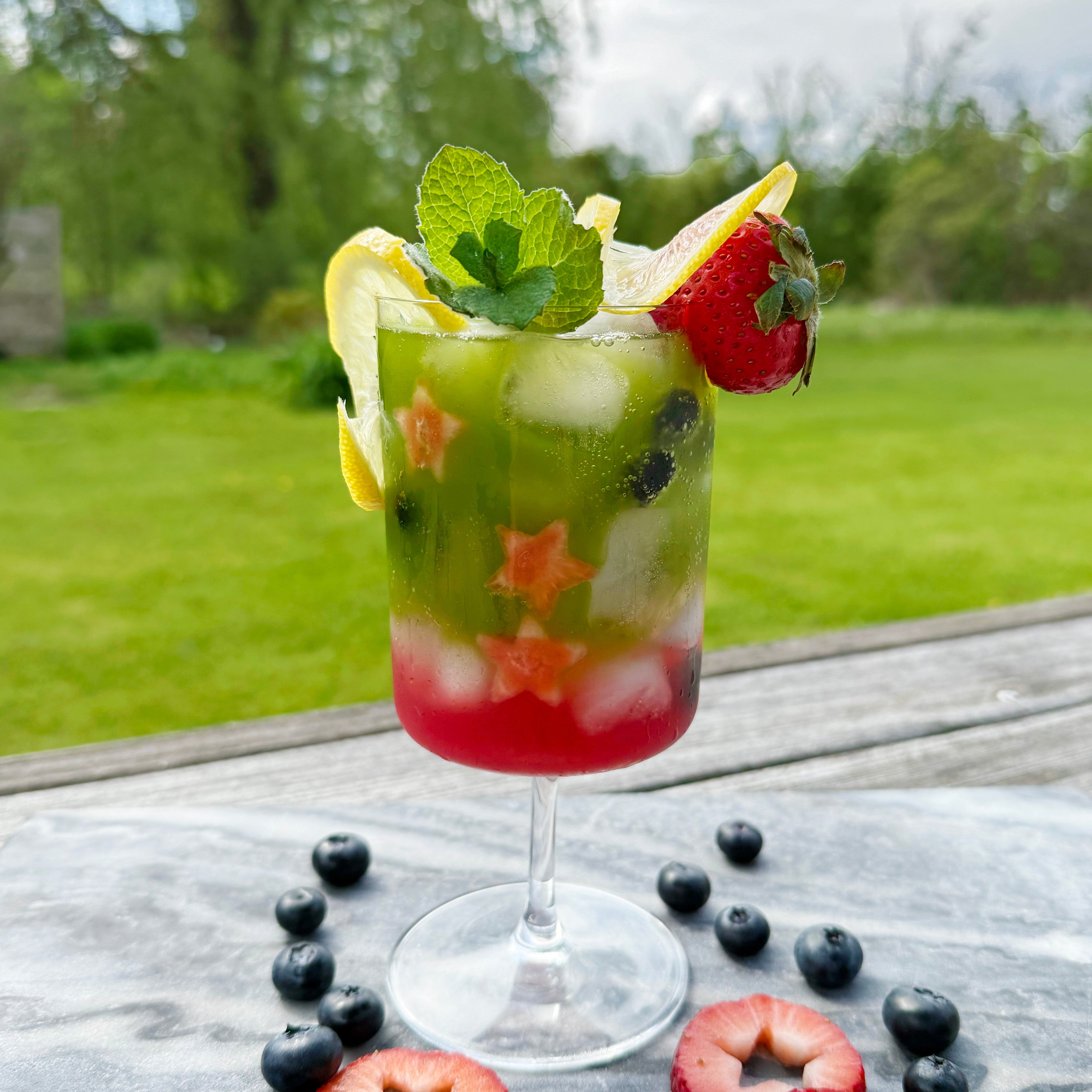 A colorful matcha lemon berry sparkler garnished with mint, lemon, and strawberry, set outdoors on a sunny day