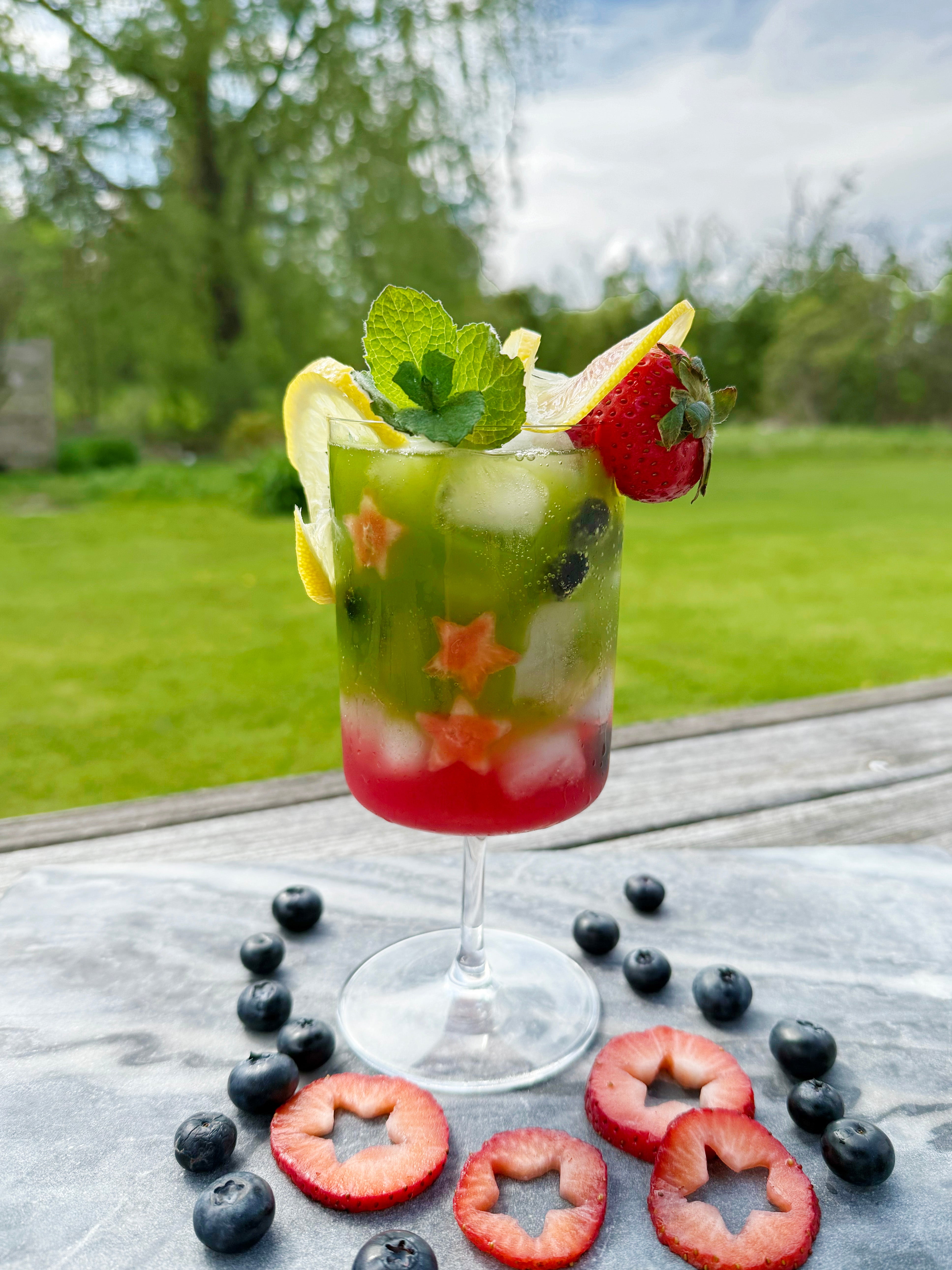 A colorful matcha lemon berry sparkler garnished with mint, lemon, and strawberry, set outdoors on a sunny day