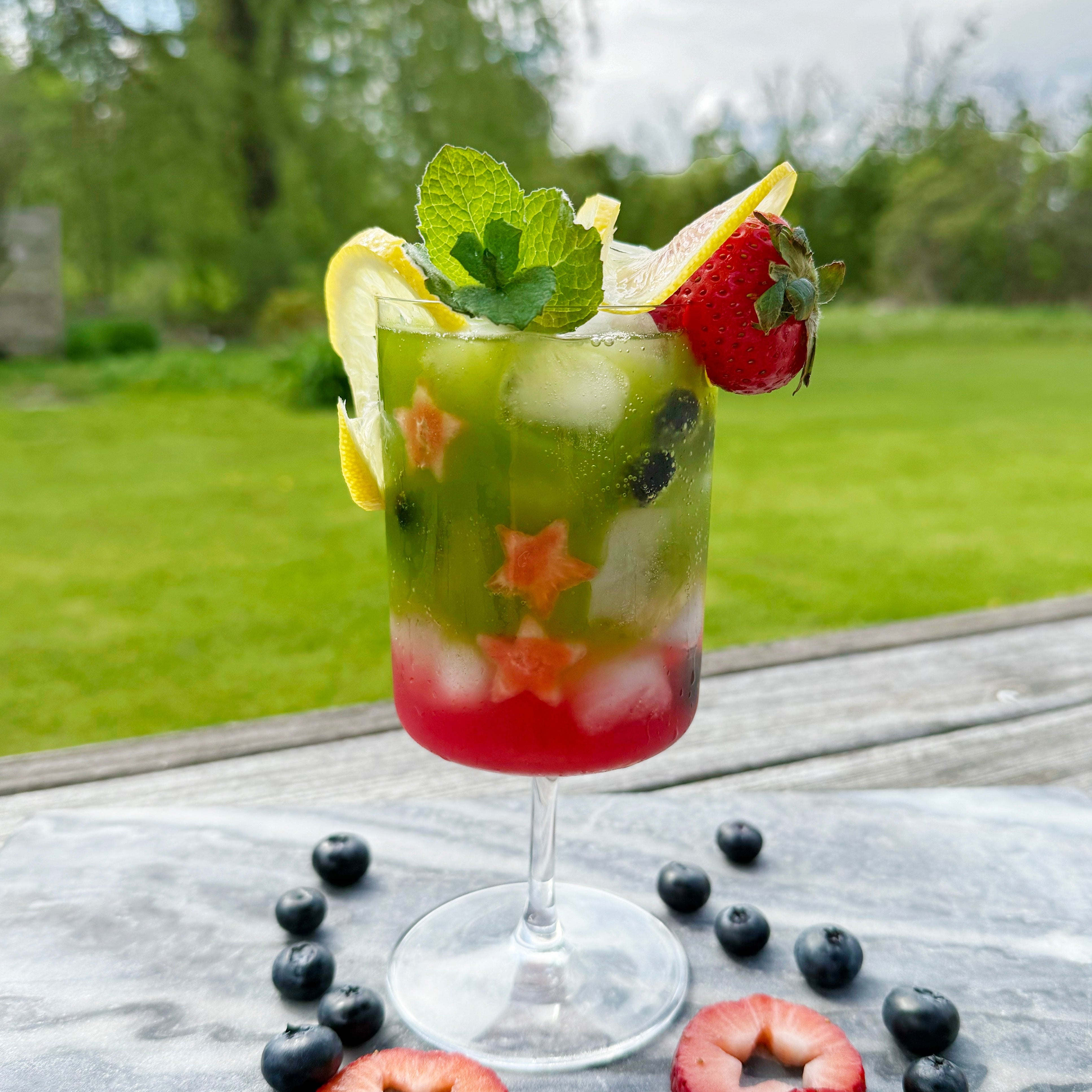 A colorful matcha lemon berry sparkler garnished with mint, lemon, and strawberry, set outdoors on a sunny day