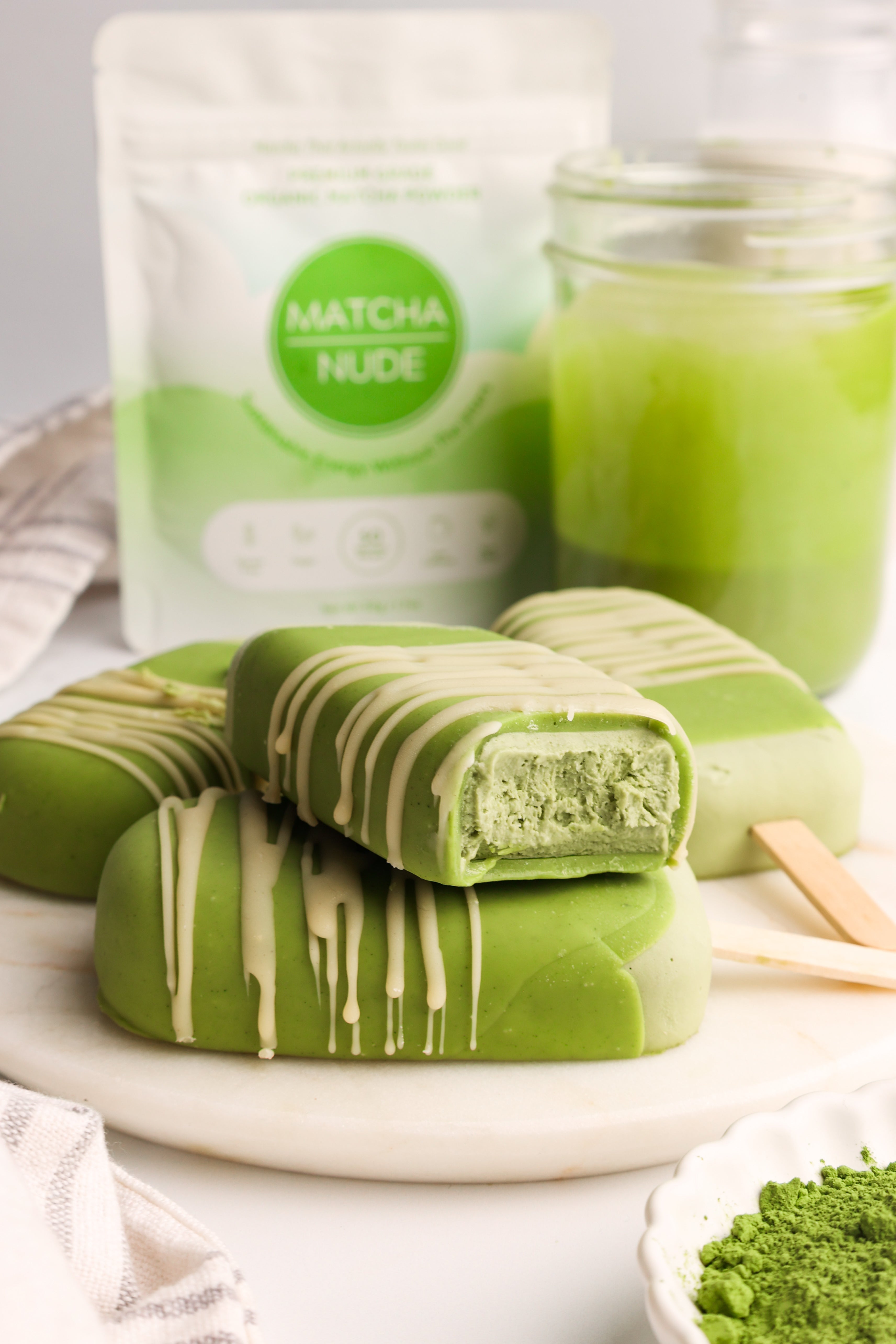 Creamy matcha ice cream bars coated in green and white matcha chocolate, stacked on a white plate with a Matcha Nude pouch and iced matcha latte in the background.