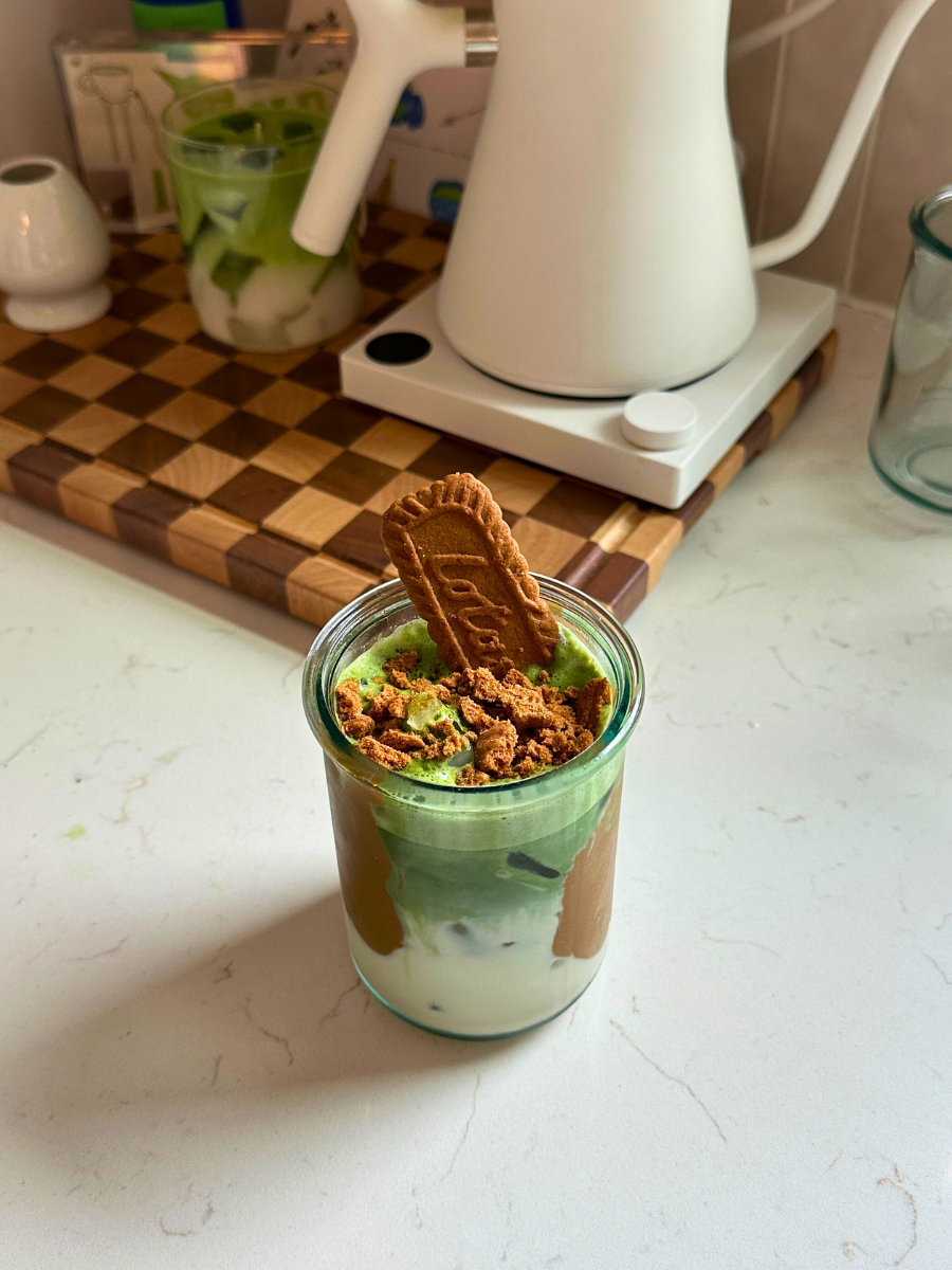Iced cookie butter matcha latte in a glass jar, topped with crushed Biscoff cookies and a whole cookie, layered with matcha and milk, set on a light kitchen counter.