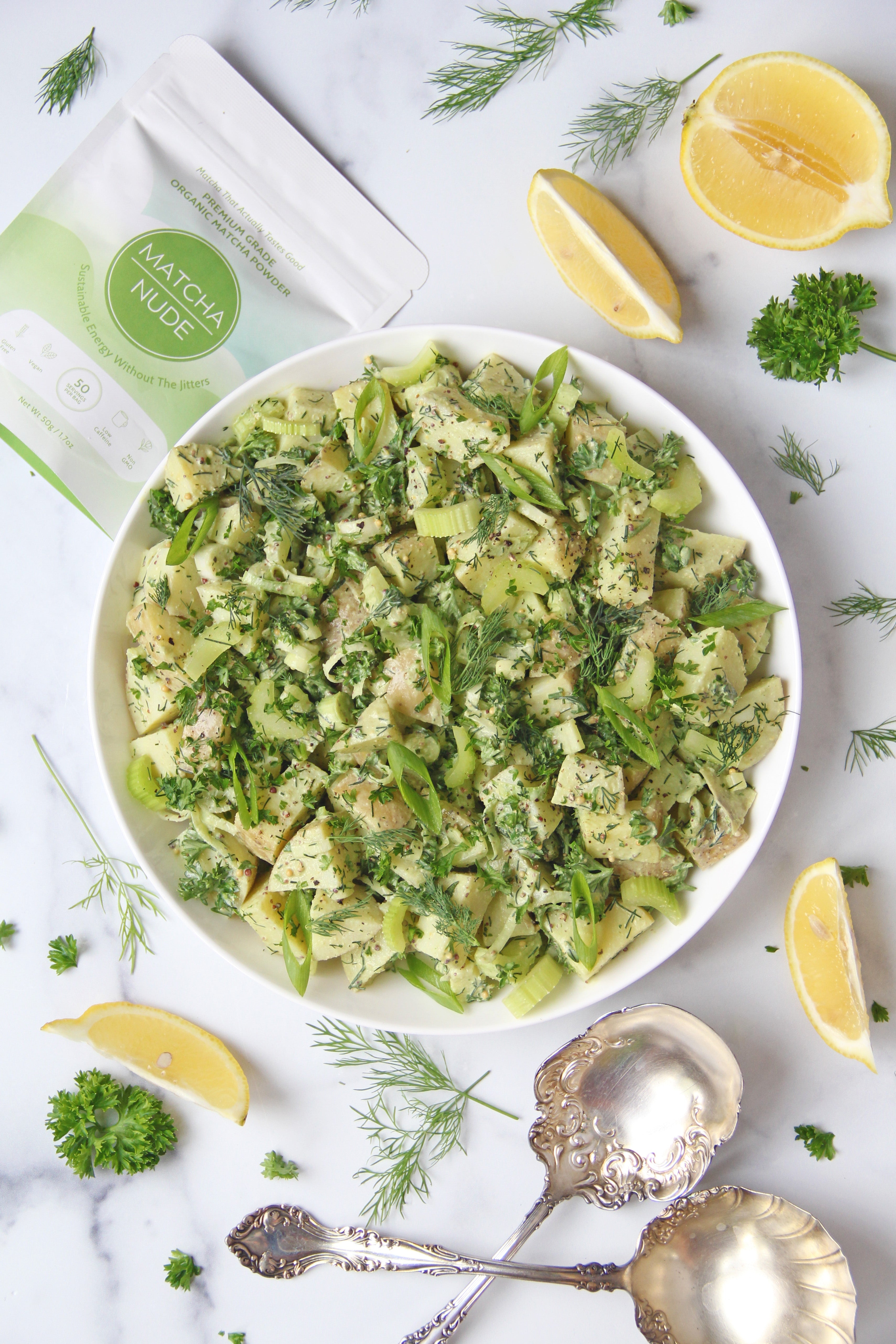 Bowl of creamy herbed potato salad made with Matcha Nude, surrounded by fresh herbs, lemon wedges, and vintage silver serving spoons on a marble counter.