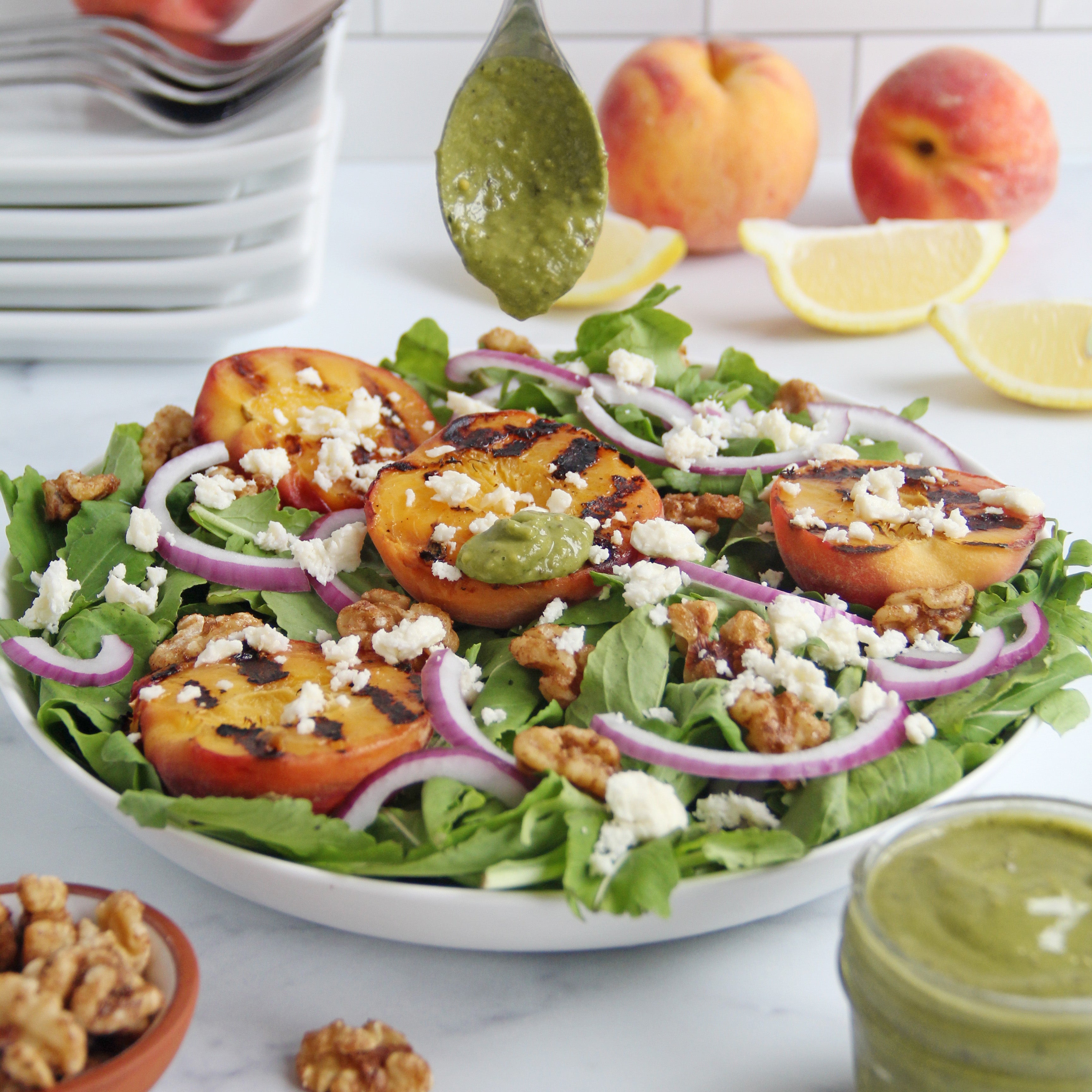 Grilled peach salad on a bed of arugula topped with red onions, feta, and walnuts, with a spoon drizzling matcha lemon vinaigrette and fresh peaches and lemon slices in the background.