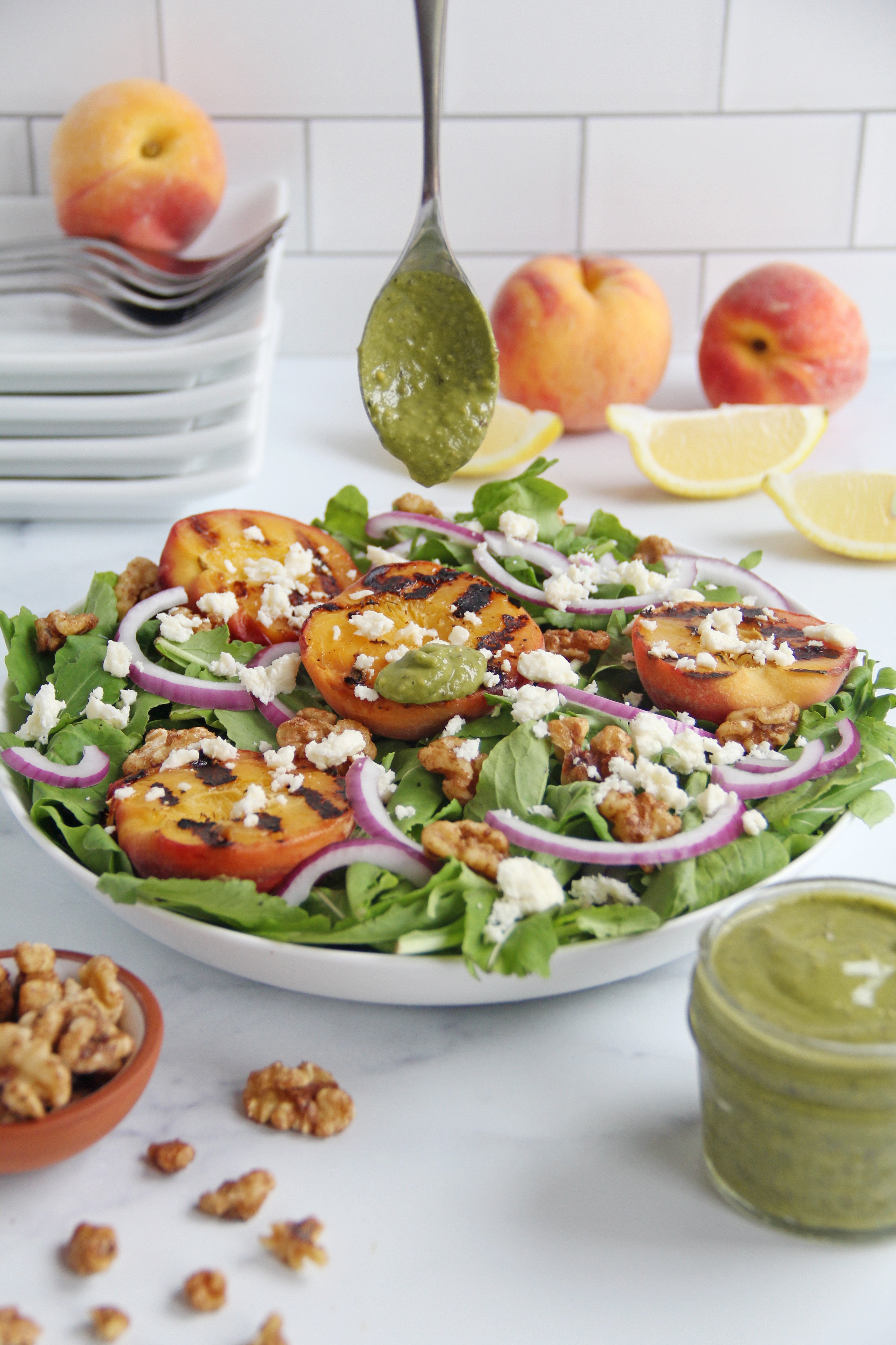 Grilled peach salad on a bed of arugula topped with red onions, feta, and walnuts, with a spoon drizzling matcha lemon vinaigrette and fresh peaches and lemon slices in the background.