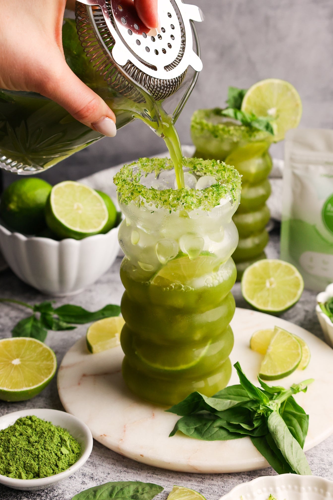 Iced matcha basil limeade being poured into a glass with a green sugar rim, surrounded by fresh limes, basil, and Matcha Nude powder.
