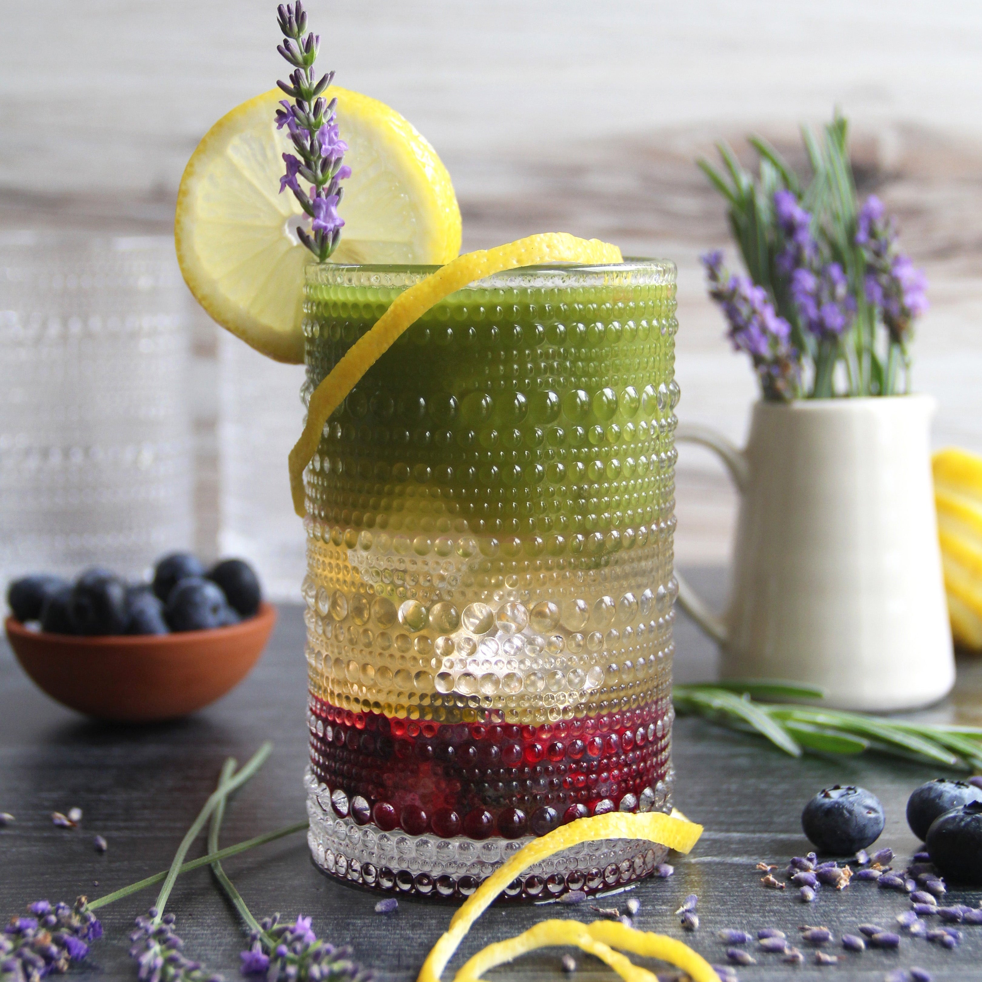 Colorful layered Matcha Lavender Lemonade Spritzer garnished with lemon slice, lavender sprig, and blueberries, served in a textured glass with fresh lavender and citrus on the table.
