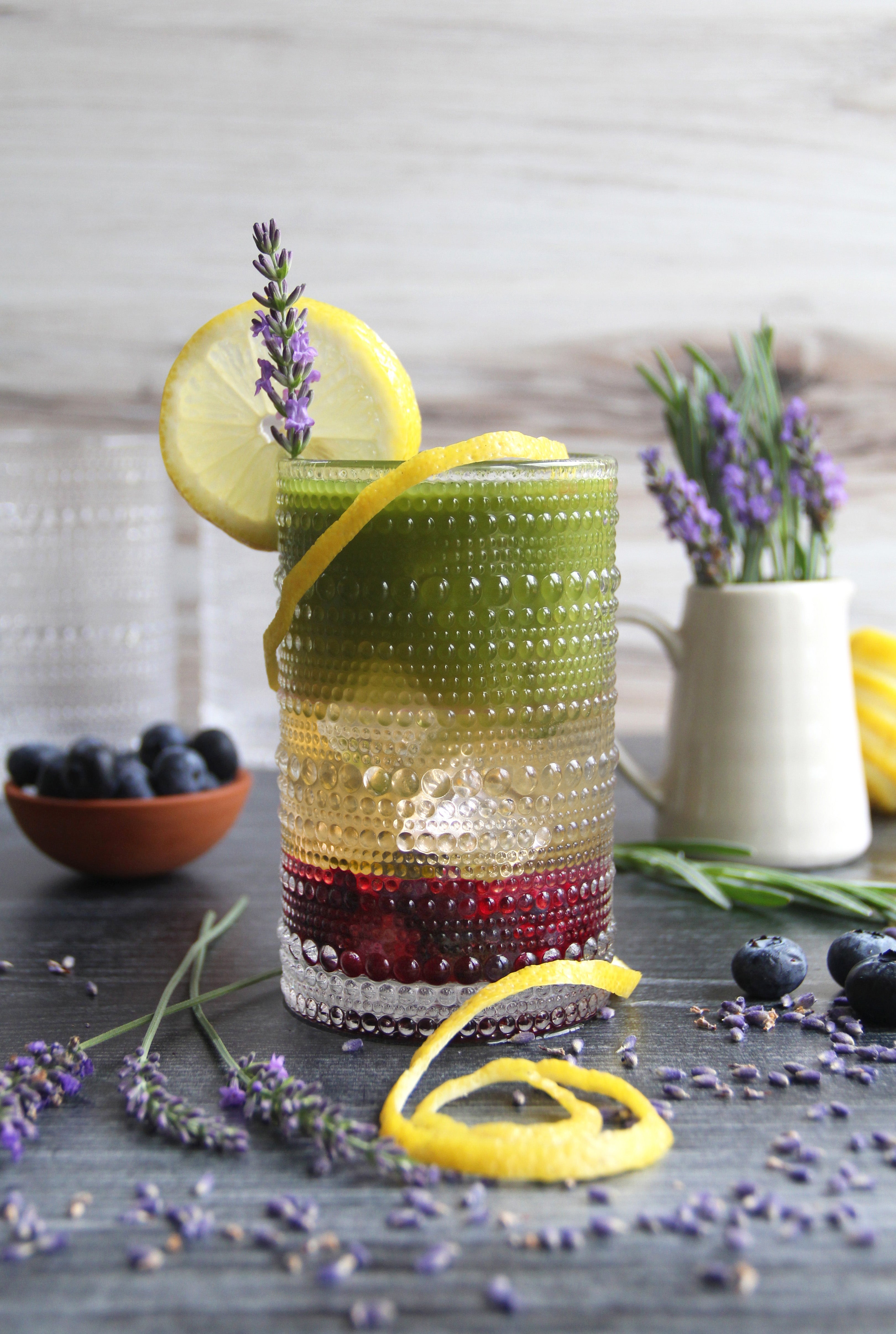 Colorful layered Matcha Lavender Lemonade Spritzer garnished with lemon slice, lavender sprig, and blueberries, served in a textured glass with fresh lavender and citrus on the table.