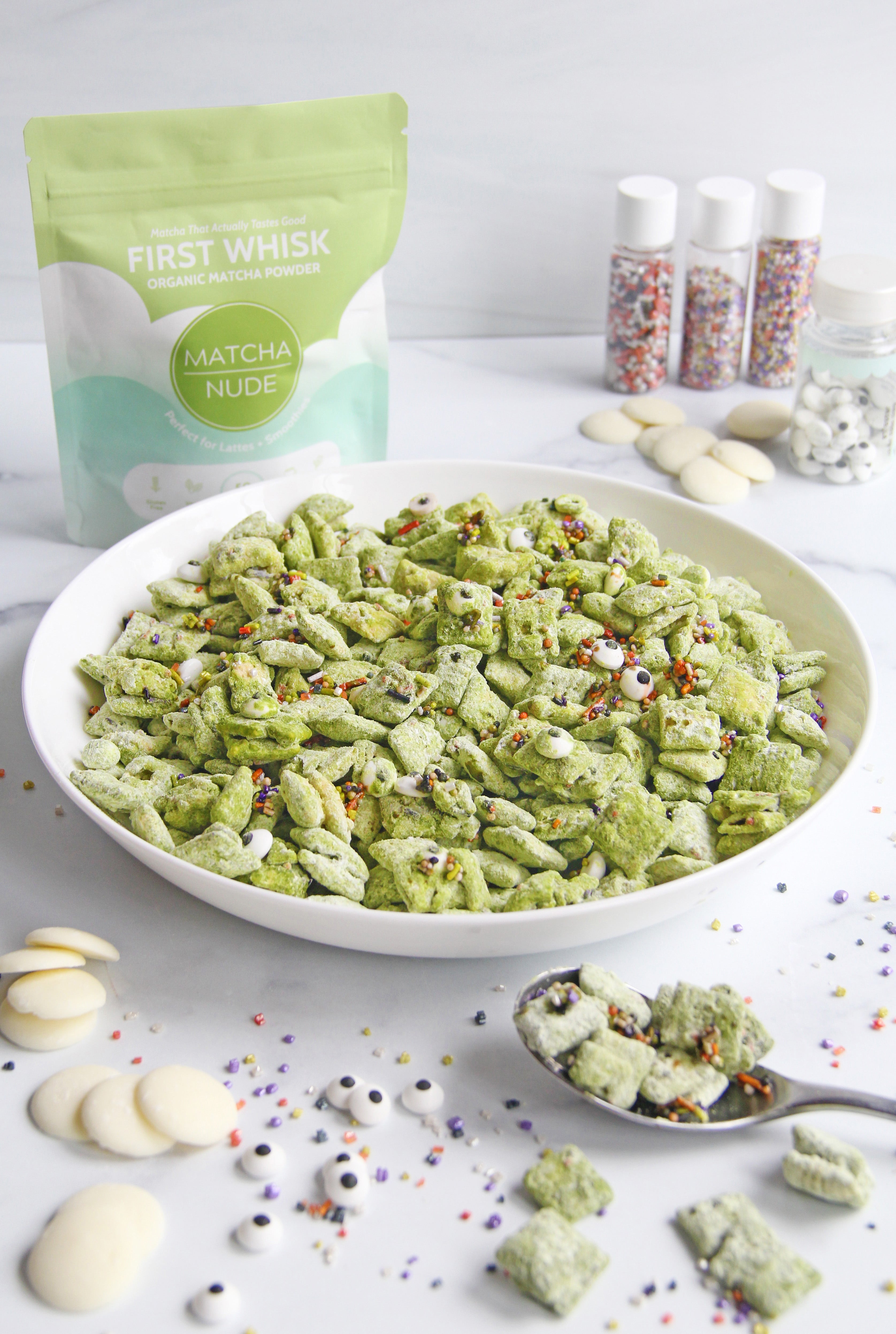 Bowl of green Monster Matcha Puppy Chow made with First Whisk, topped with Halloween sprinkles and candy eyes, surrounded by white chocolate and decorations.