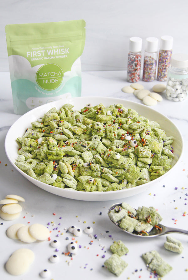 Bowl of green Monster Matcha Puppy Chow made with First Whisk, topped with Halloween sprinkles and candy eyes, surrounded by white chocolate and decorations.