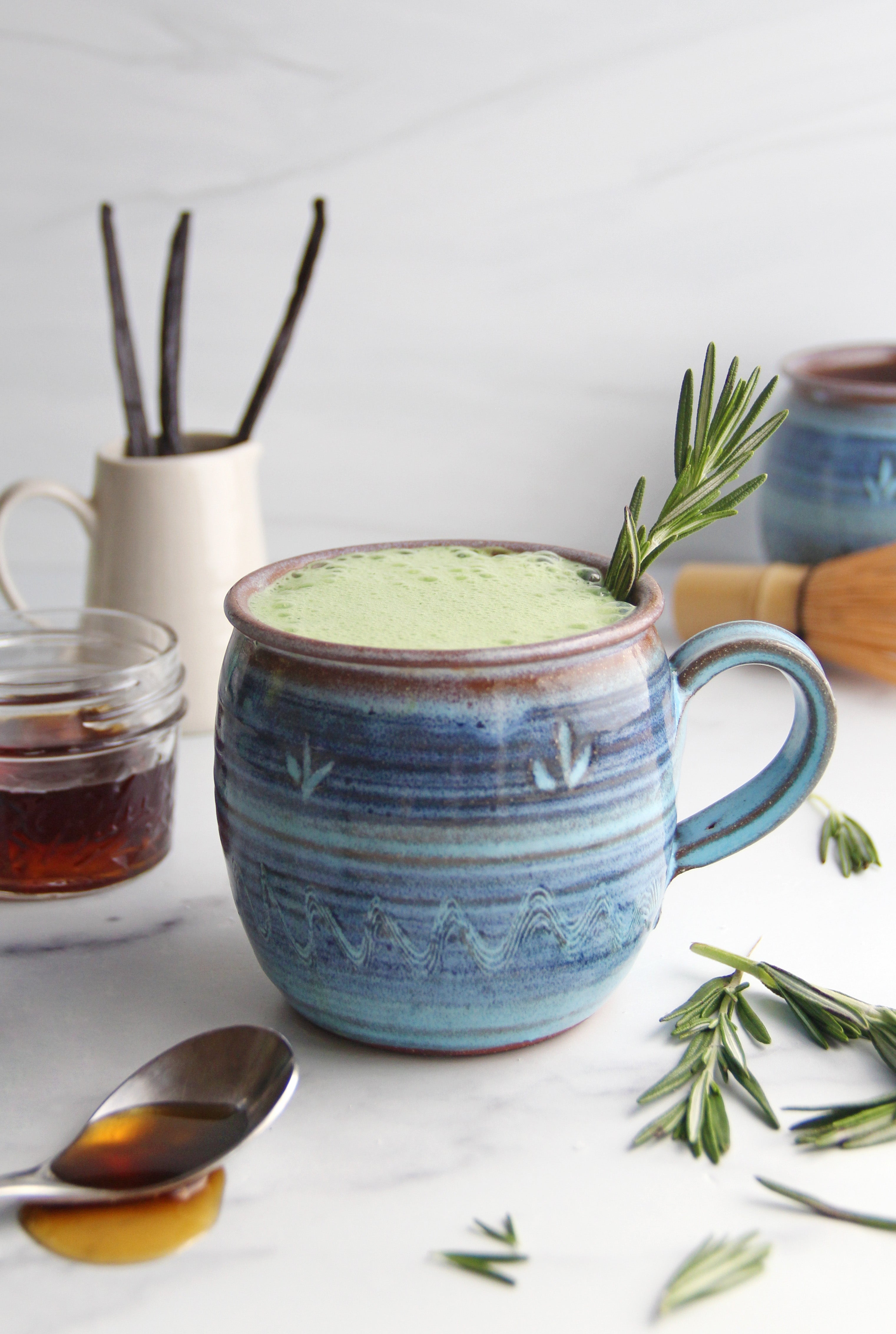 Rosemary Vanilla Matcha Latte in a handmade ceramic mug, garnished with fresh rosemary and surrounded by vanilla beans, maple syrup, and matcha tools.