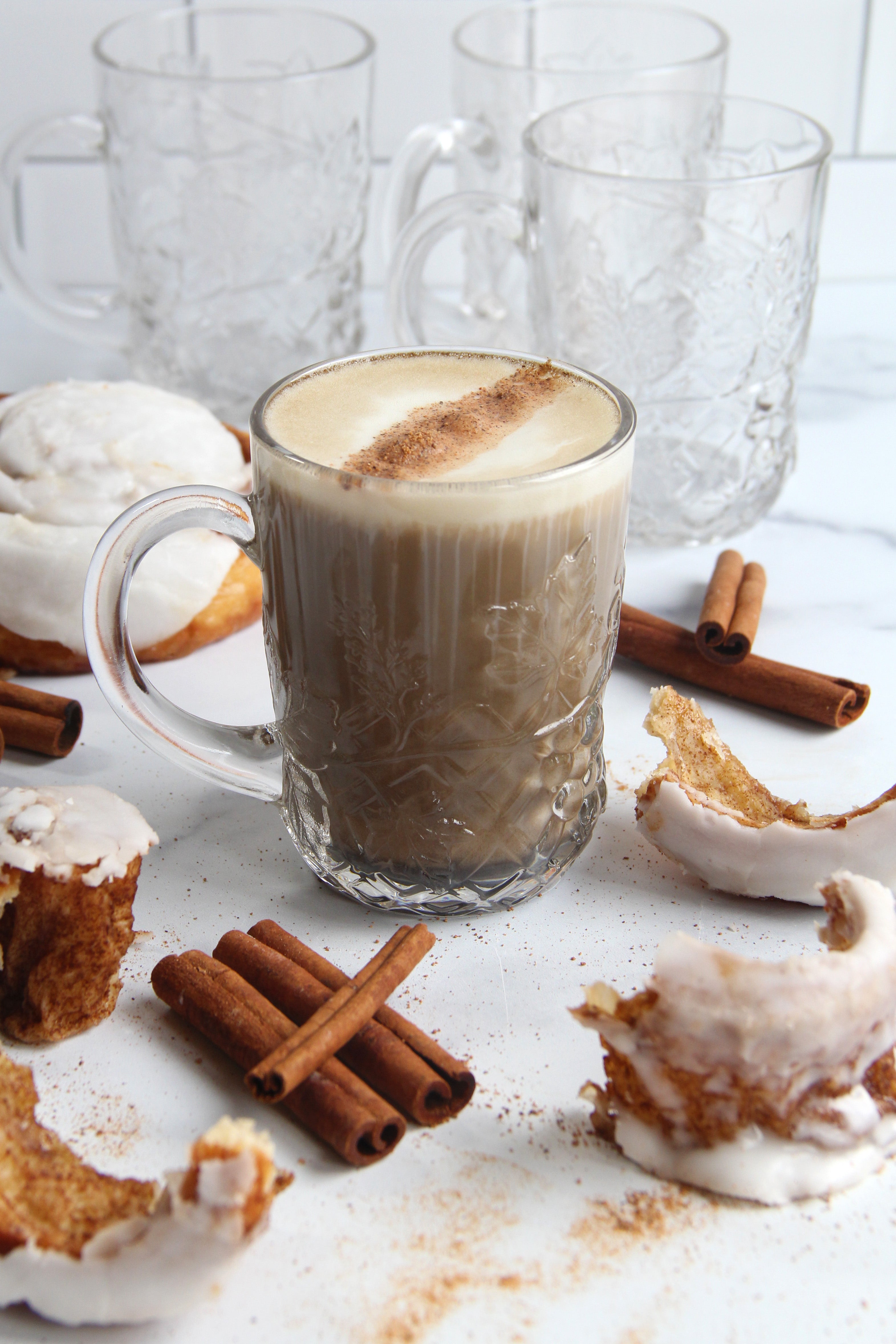 Glass mug of Cinnamon Roll Hojicha Latte topped with cinnamon, surrounded by cinnamon sticks and frosted cinnamon rolls on a white background.
