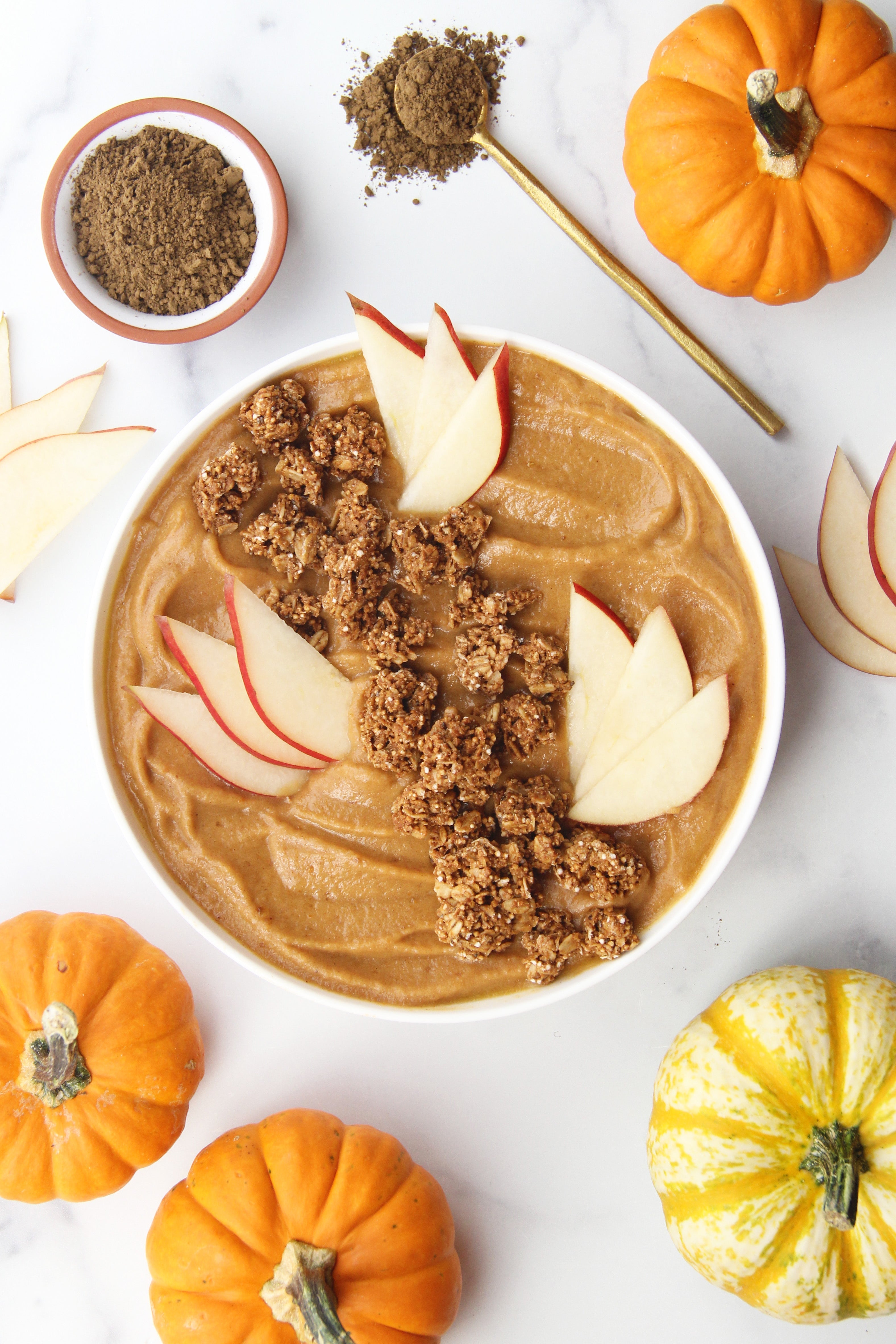 Bowl of pumpkin spice hojicha smoothie topped with red pear slices and granola, surrounded by pumpkins and roasted tea powder on a white background.
