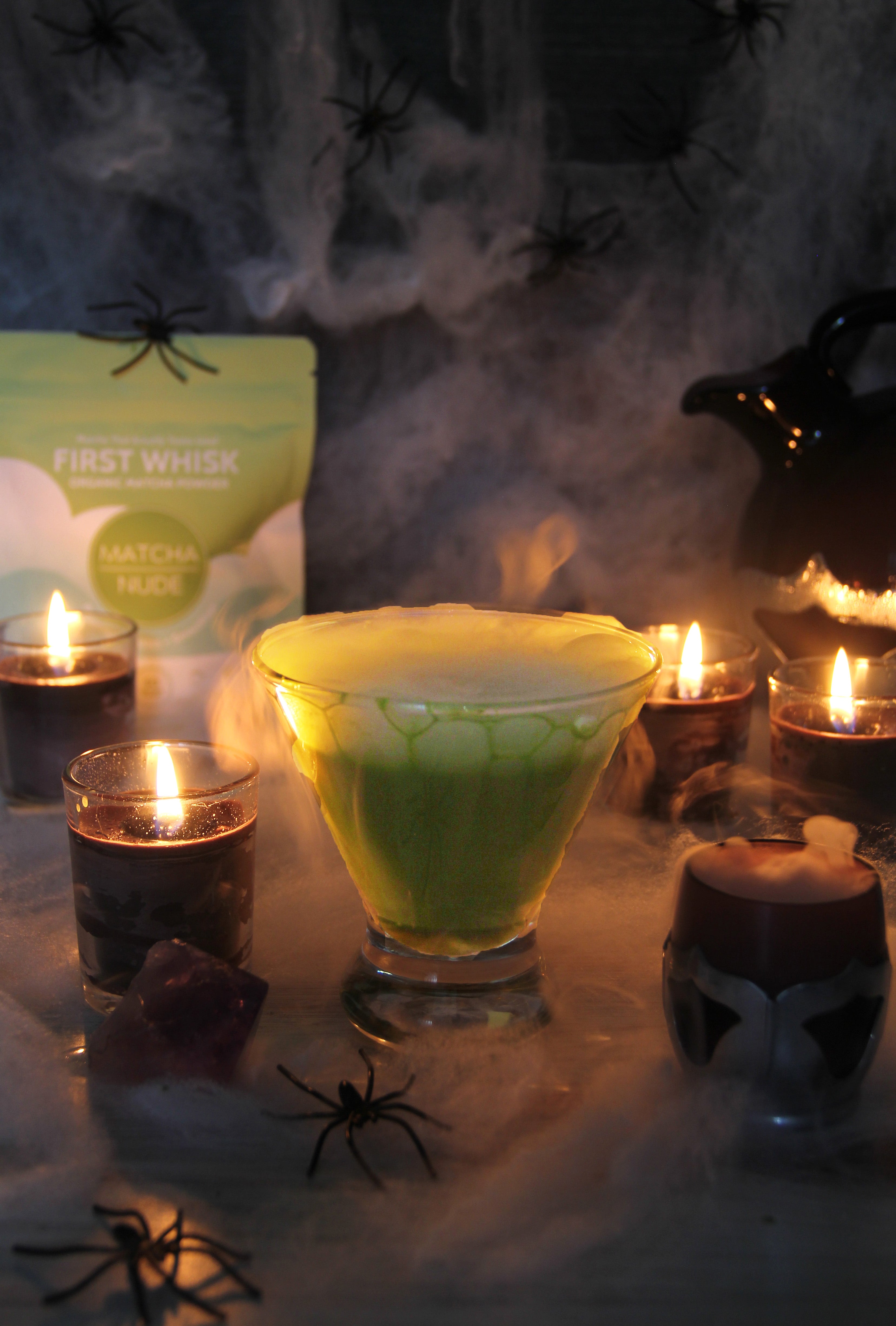 Green FrankenFizz matcha cocktail surrounded by candles, cobwebs, and fake spiders with Matcha Nude First Whisk pouch in the background