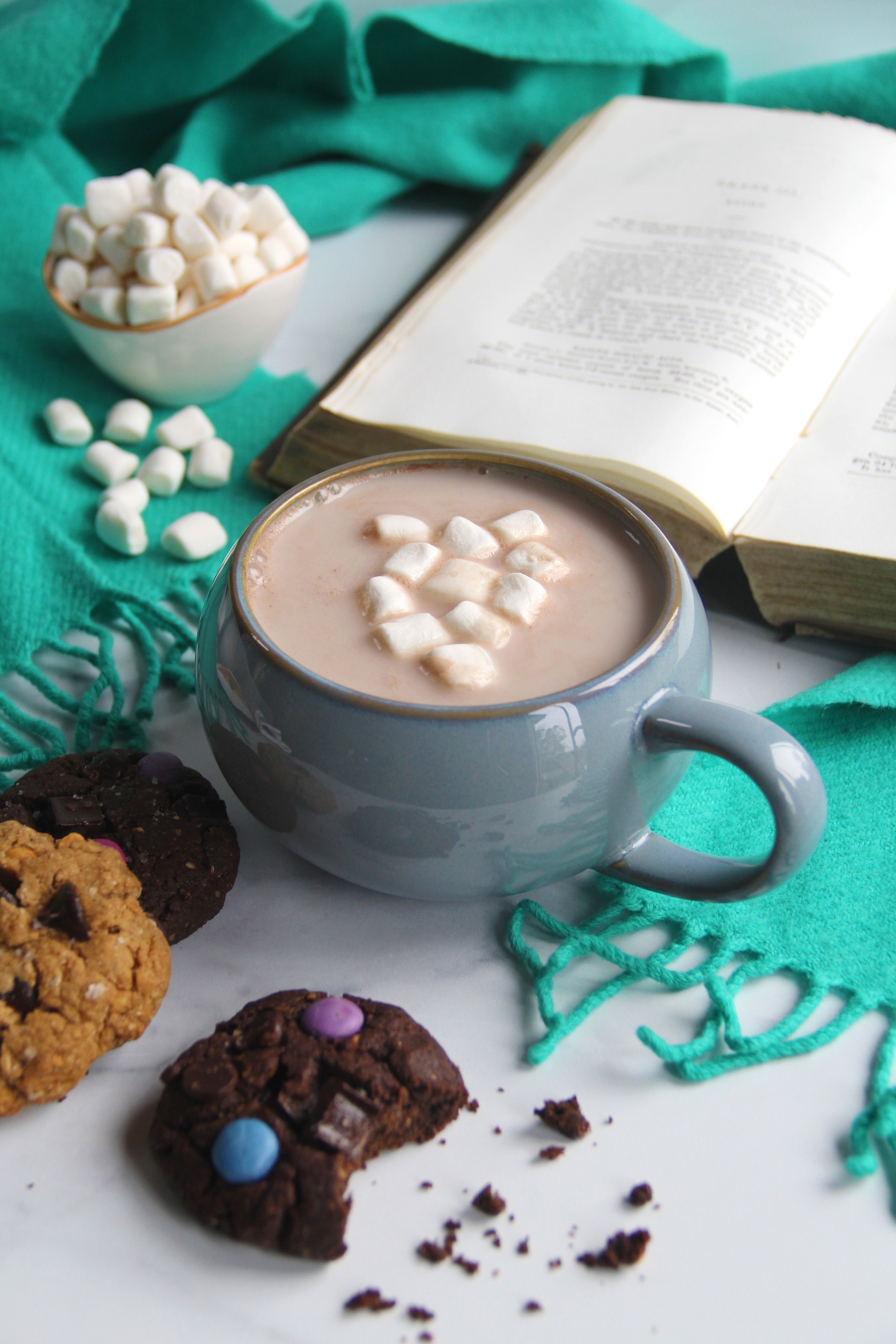 A cozy mug of hojicha hot chocolate topped with mini marshmallows, surrounded by colorful cookies, a bowl of marshmallows, and an open book on a teal blanket.