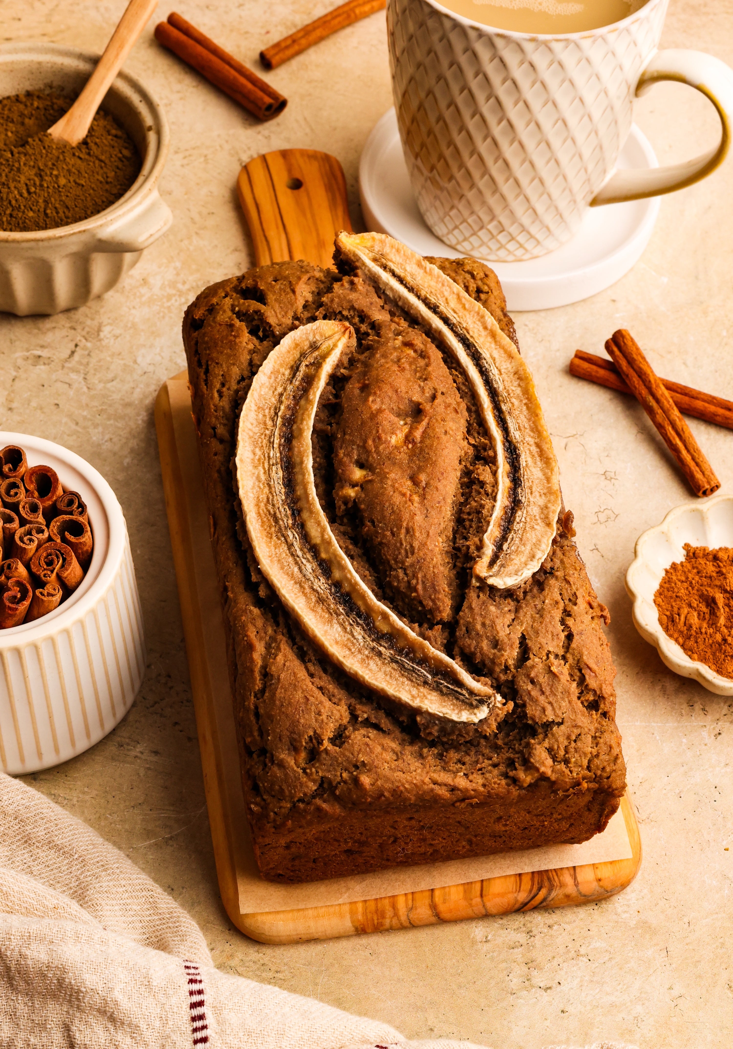 A golden brown loaf of hojicha banana bread topped with sliced banana, surrounded by a mug of tea, cinnamon sticks, and a dish of hojicha powder.