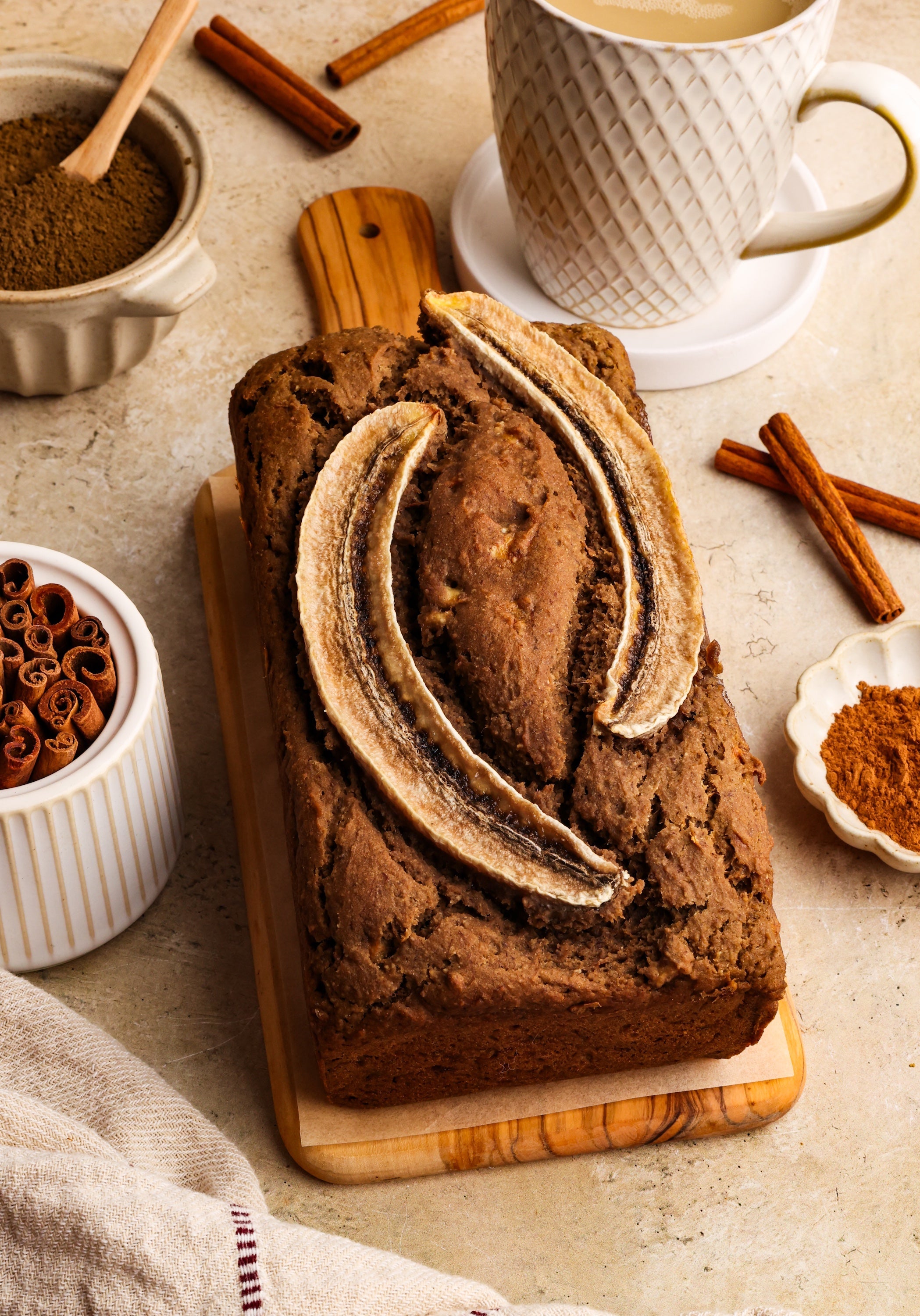 A golden brown loaf of hojicha banana bread topped with sliced banana, surrounded by a mug of tea, cinnamon sticks, and a dish of hojicha powder.