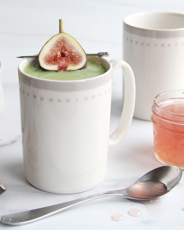 A white mug of matcha latte topped with a fig half, surrounded by pink fig honey syrup, a spoon, and a soft pastel aesthetic, perfect cozy vibes.