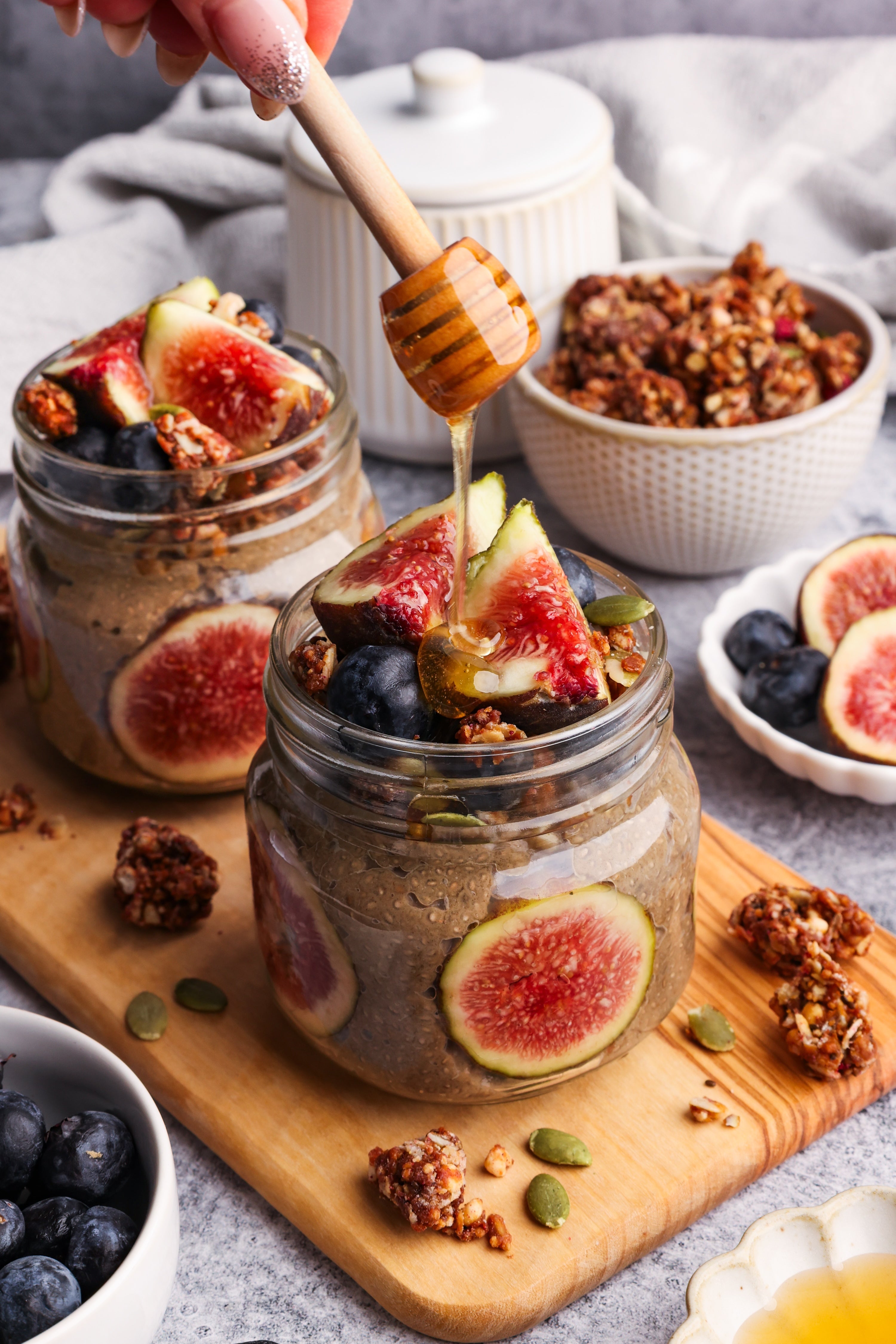 Jar of hojicha chia seed pudding topped with fresh figs, blueberries, granola, and honey drizzle, styled on a wooden board with additional fruit and granola bowls in the background.