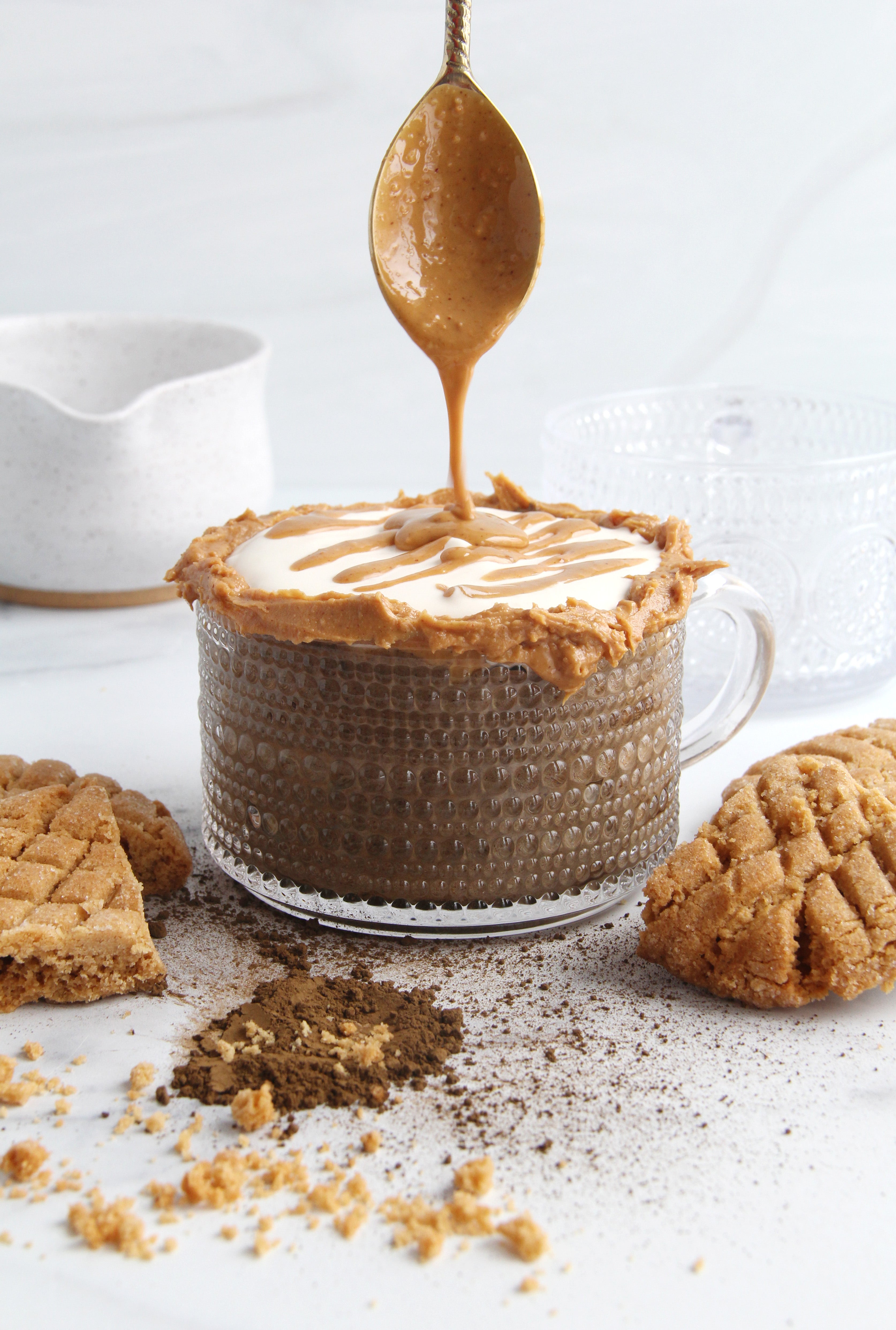 Glass mug of peanut butter hojicha latte topped with creamy foam and a peanut butter drizzle, surrounded by cookie crumbles and hojicha powder on a white surface.
