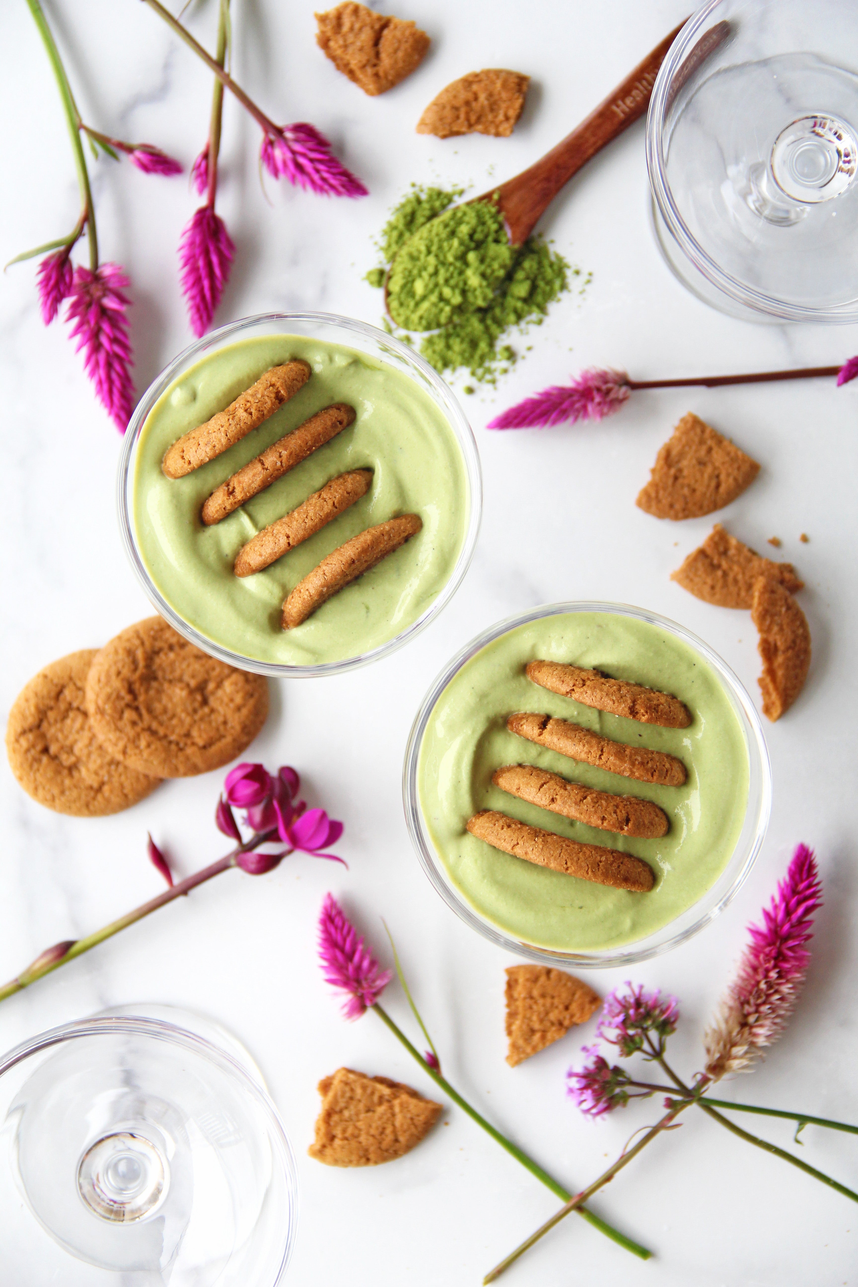 Two matcha coconut yogurt cheesecakes in glass dessert cups, topped with gingersnap cookies. Surrounded by bright pink flowers, cookie crumbs, and a wooden spoon with vibrant matcha powder.