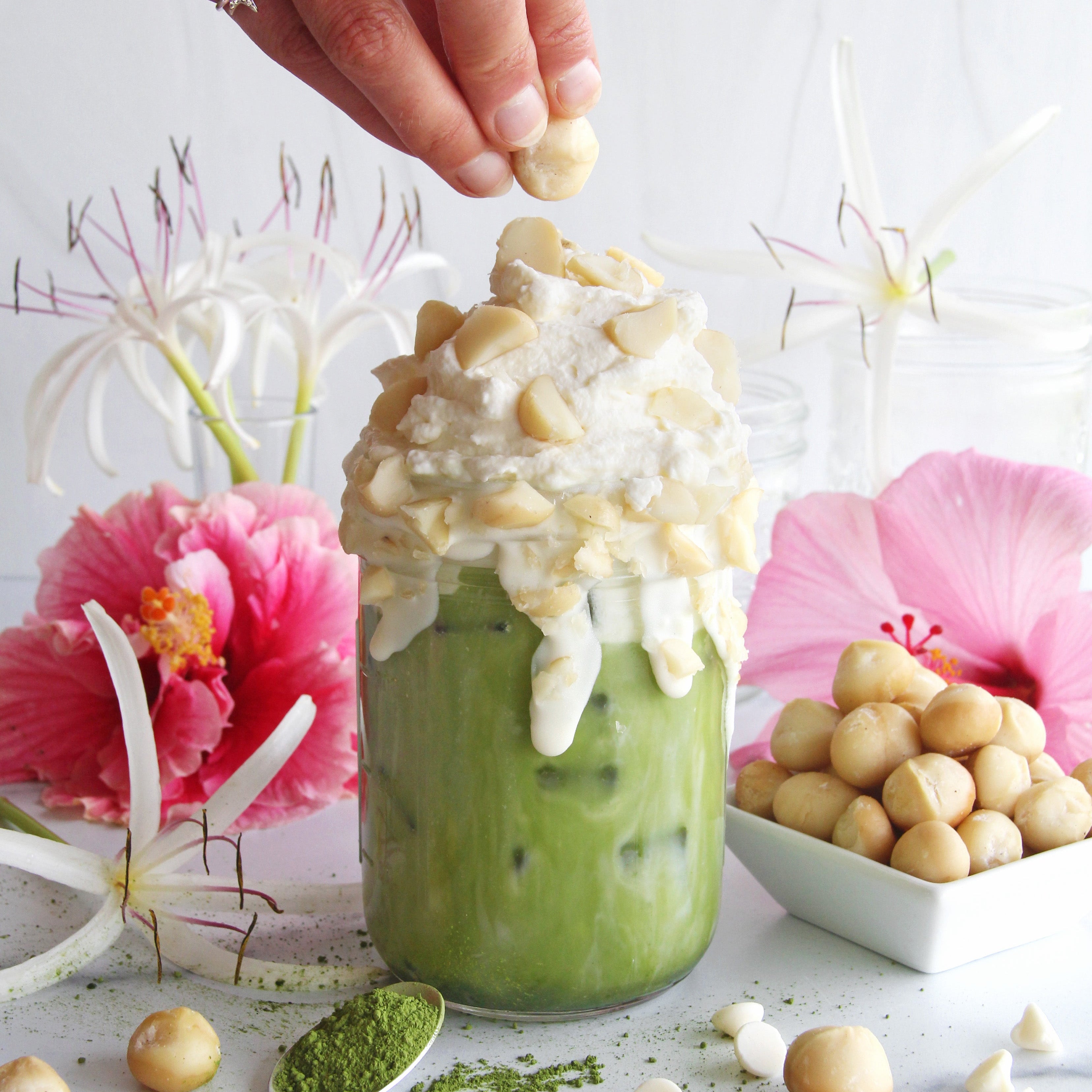 Iced matcha latte topped with whipped cream and macadamia nuts, surrounded by white chocolate chips, matcha powder, and pink hibiscus flowers.