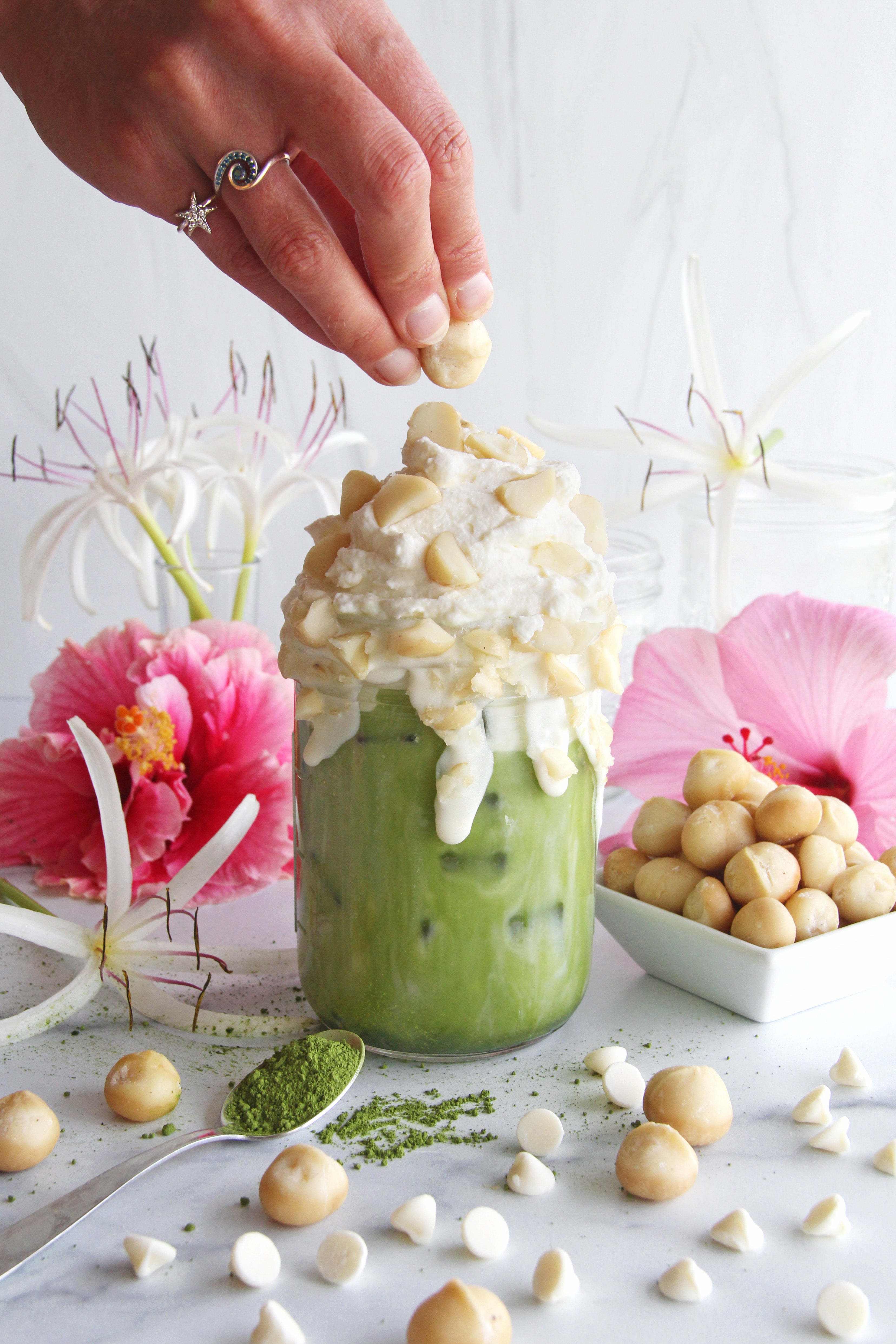 Iced matcha latte topped with whipped cream and macadamia nuts, surrounded by white chocolate chips, matcha powder, and pink hibiscus flowers.