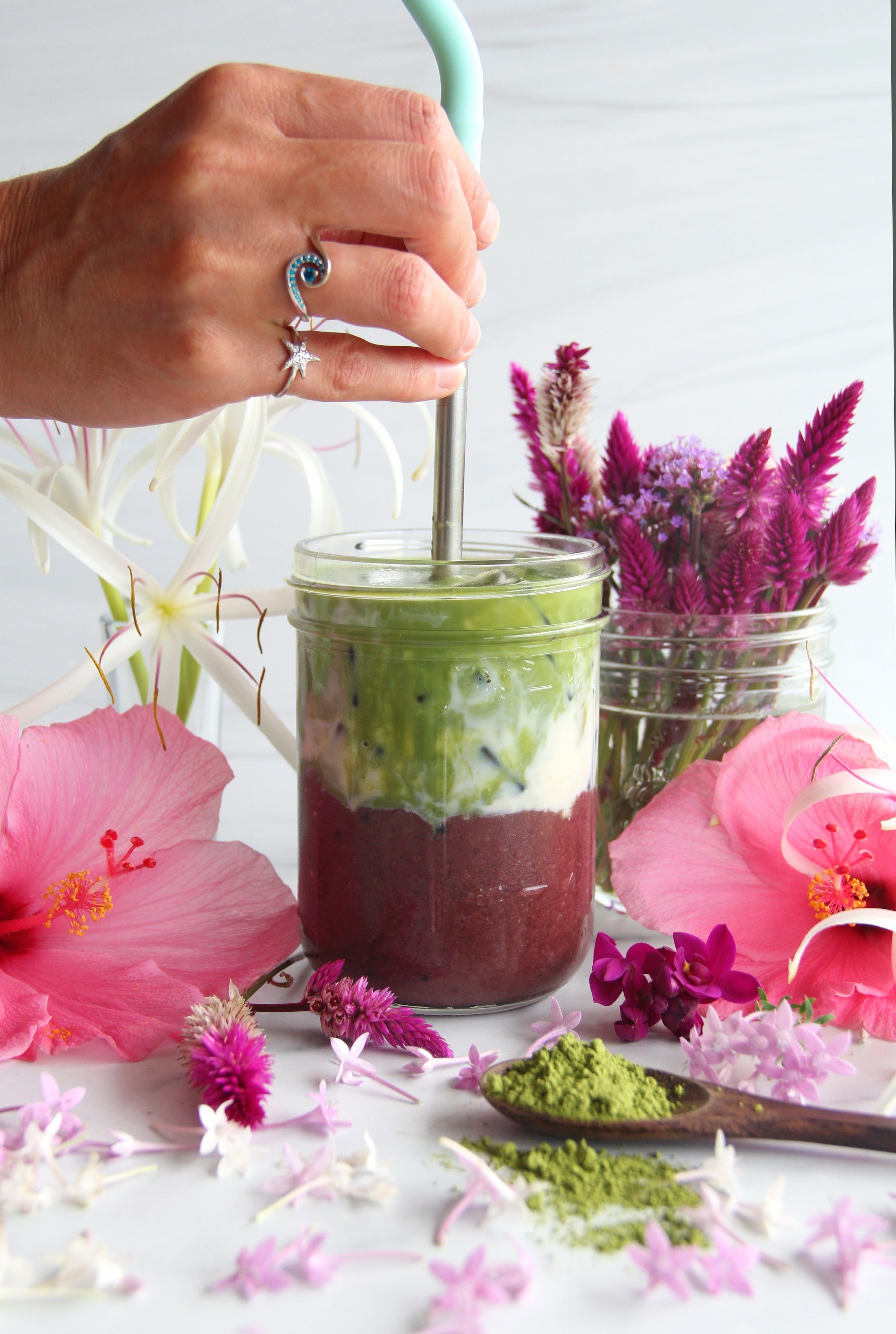 Hand stirring layered iced açaí matcha latte in a jar, surrounded by pink flowers and matcha powder.