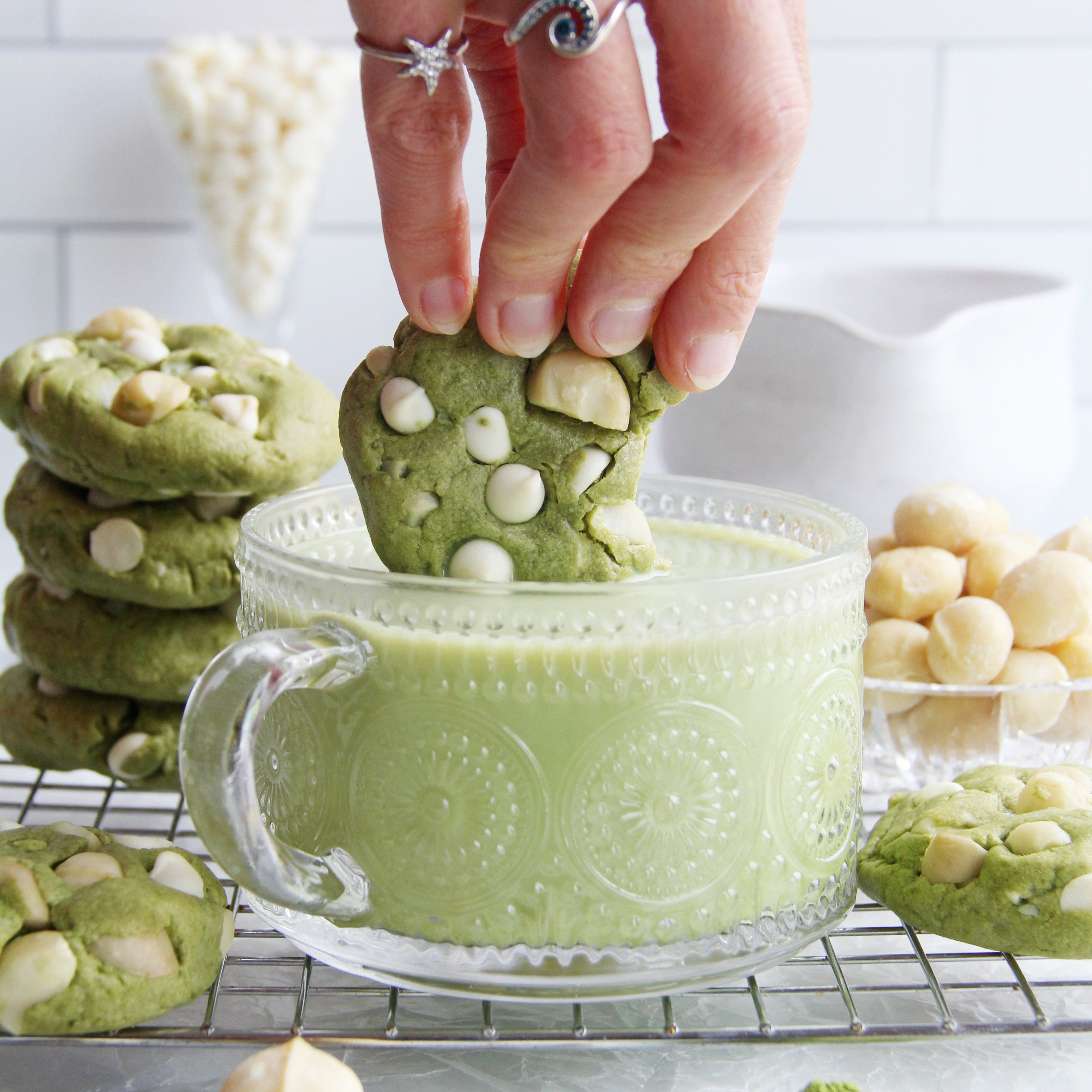 Matcha white chocolate macadamia cookie dipped into a glass of matcha latte with cookies and ingredients around.