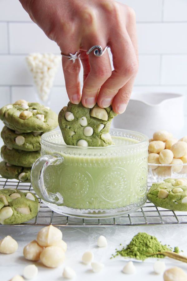 Matcha white chocolate macadamia cookie dipped into a glass of matcha latte with cookies and ingredients around.