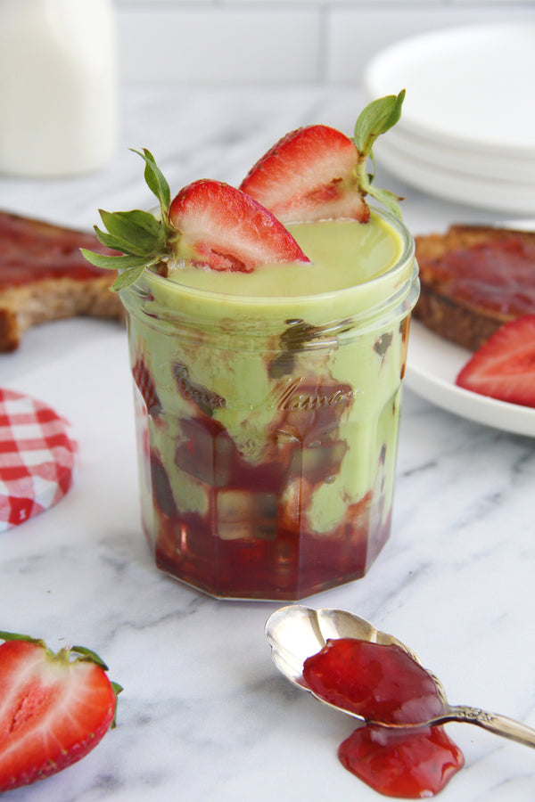 Grandma's Jam Jar matcha latte in a glass mason jar with strawberry jam layered on the bottom, topped with sliced strawberries, surrounded by toast and fresh strawberries on a marble countertop