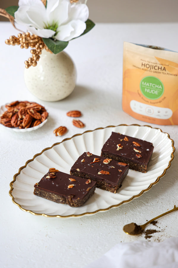 Hojicha no-bake brownies topped with a rich chocolate layer, chopped pecans, and flaky sea salt, served on a white scalloped platter beside a bag of Matcha Nude hojicha.