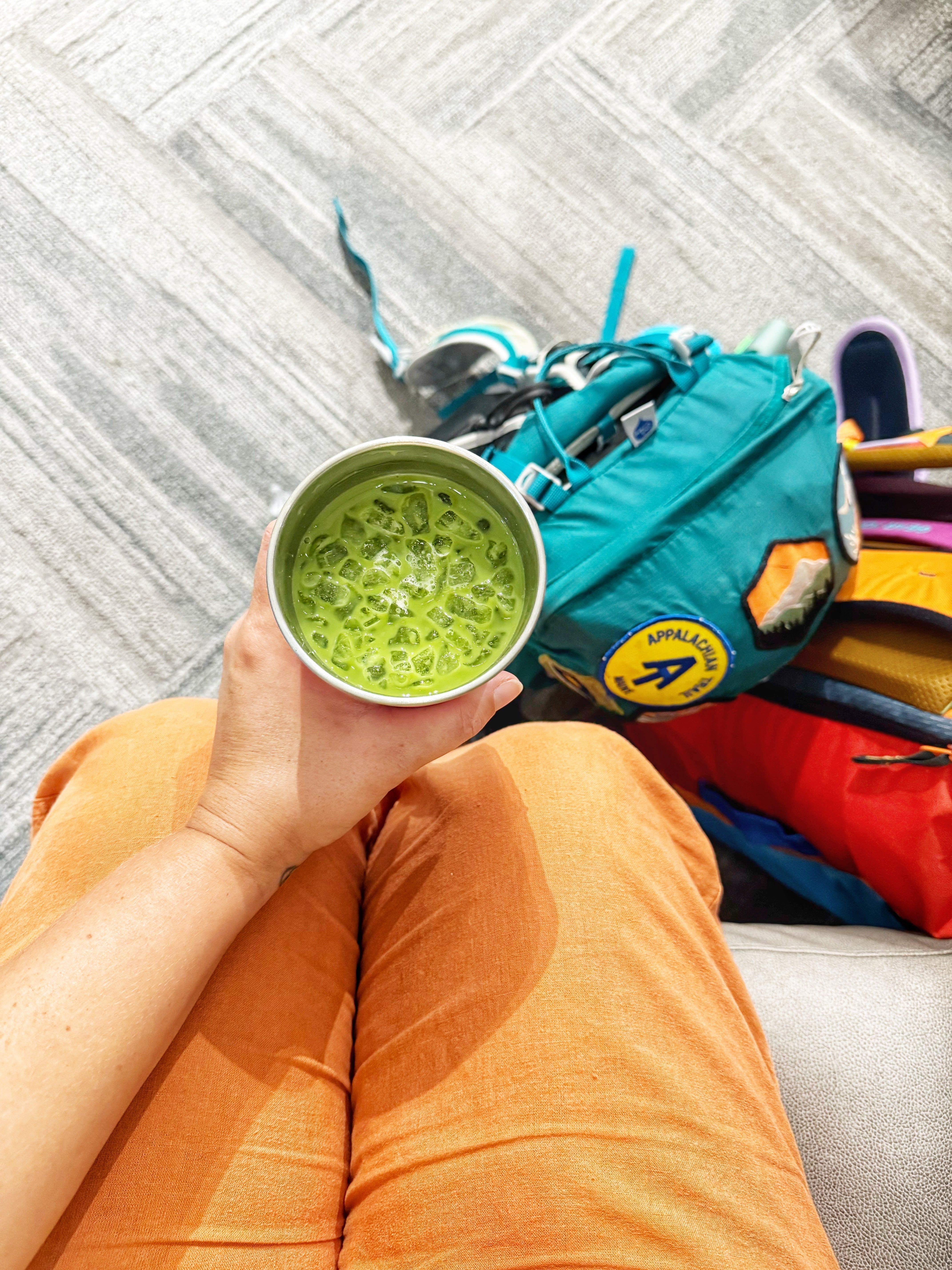 Iced matcha in hand with backpack and travel gear nearby, cozy on-the-go moment.