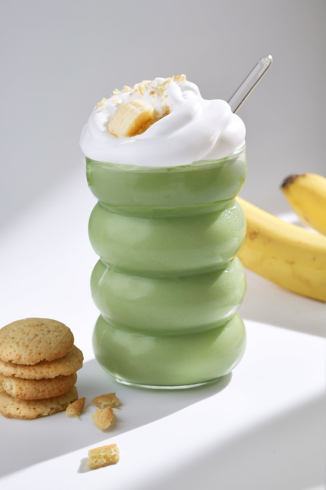 A vibrant green banana pudding matcha latte topped with a generous swirl of whipped cream and banana slices, paired with a stack of golden nilla wafer cookies and fresh bananas in the background.