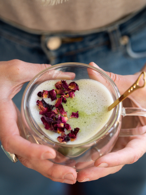 Matcha Latte in a heart shaped glass mug with rose petals and a gold spoon held by two hands