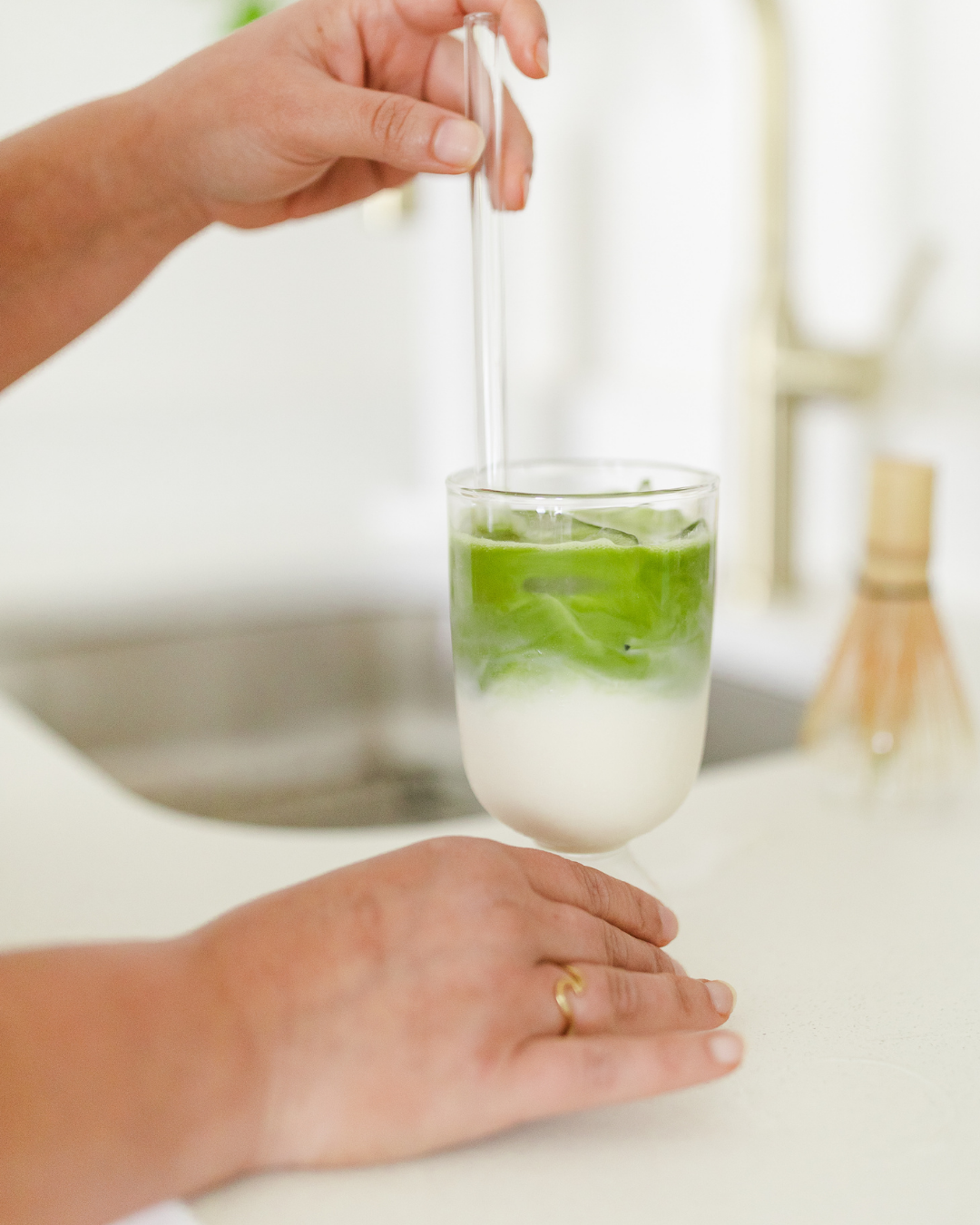 Women's hands mixing an iced matcha latte on a kitchen counter with matcha whisk in background