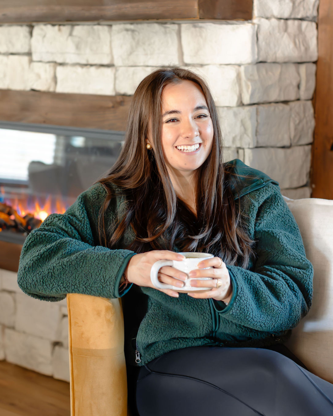 women holding a warm mug of matcha wearing a green fleece on a cozy chair near a fireplace